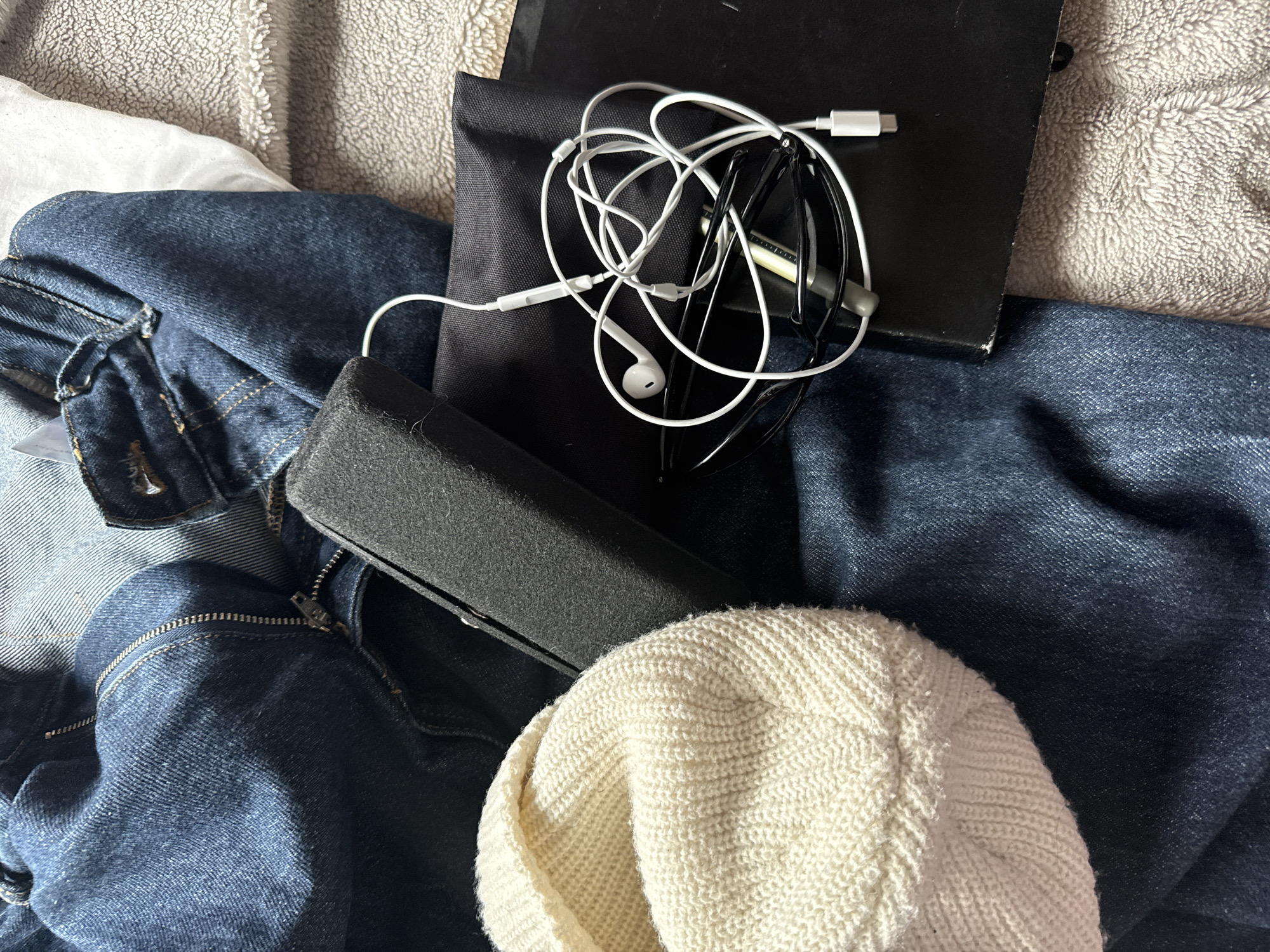 The image shows a cluttered scene with various items. A denim jacket is visible in the lower left corner of the frame. A white knit beanie rests partially on the denim jacket. A dark-colored, rectangular object that resembles a glasses case sits above the beanie. A black pouch with a soft texture is positioned above the glasses case and denim. On top of the black pouch, there is a tangled set of white wired earphones and a pair of black framed glasses. A black notebook is partially visible in the upper right corner of the frame, sitting next to the pouch. The background consists of a textured blanket or bedspread. The location is Unknown, Unknown.