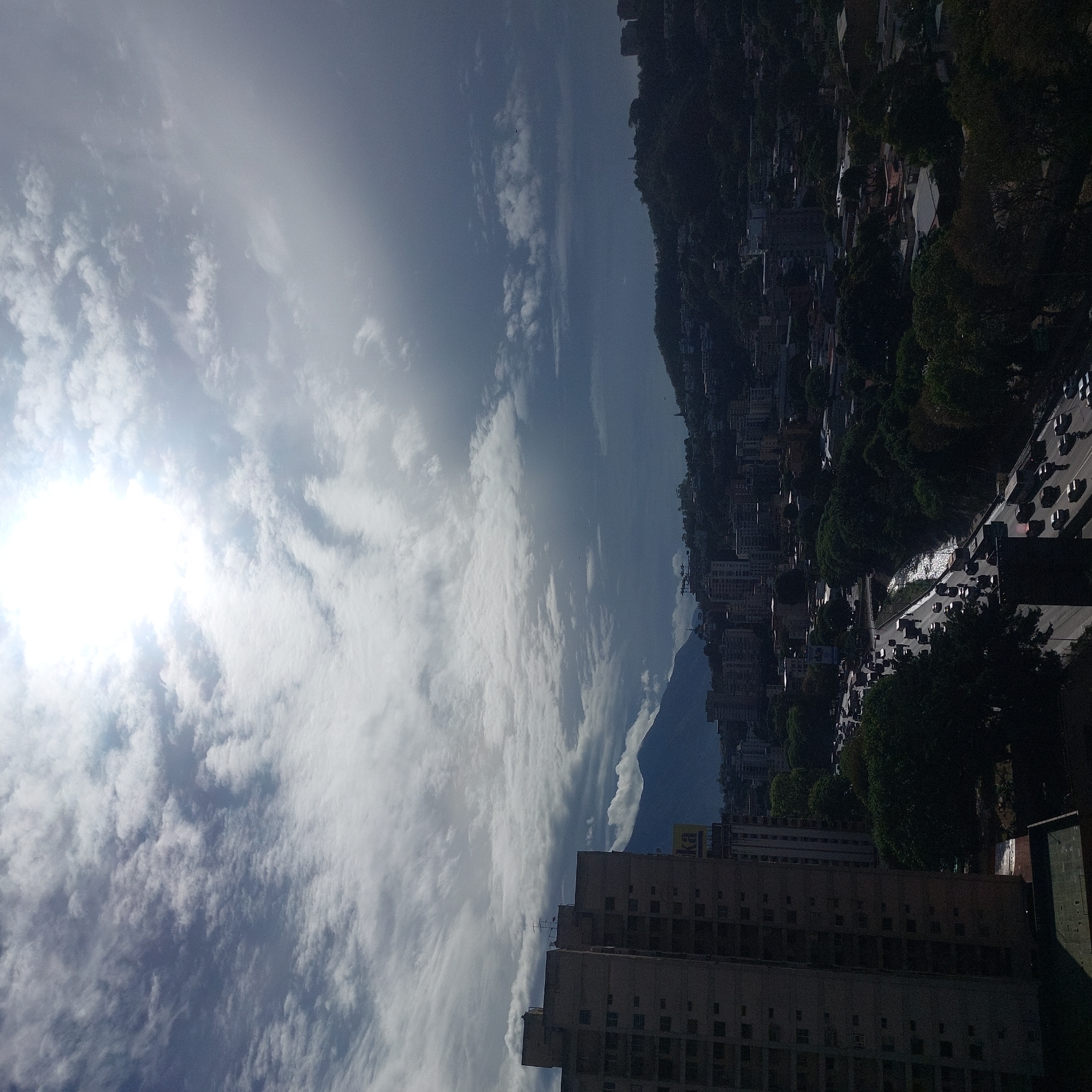 The image shows a cityscape of Caracas, Venezuela, under a sky with abundant white clouds and a bright sun. A large building dominates the foreground. In the distance, a mountain silhouette is visible. A line of trees and buildings runs along the right side of the image, appearing to border a road with active traffic. The city extends toward the base of the mountain, with buildings scattered across the hillside.