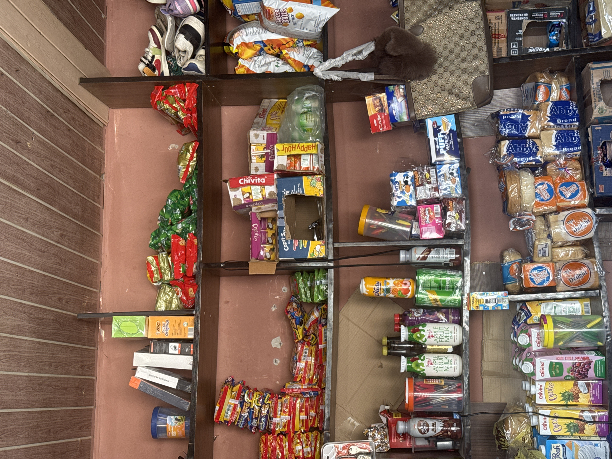 This is a photograph of a small convenience store shelf stocked with various goods. The shelf is made of dark wood and the wall behind it is painted a reddish-pink color. The shelves are organized with packaged snacks, drinks, and other items.
On the top shelf, there are several bags of chips and some footwear, possibly shoes or sandals. Below that, the shelves are filled with a variety of items including juice boxes (Chivita), bottles of juice or milk, and packaged bread. There are also some electronic items like a wireless headset.
On the right side of the shelves, more packaged bread loaves are visible, along with various drink bottles and cartons. The overall impression is of a well-stocked but somewhat cluttered shop. The lighting suggests it is daytime, and the setting appears to be an indoor retail space. There is visible text on many of the products, including brand names like "Chivita," "BIC," "Peak," and "Duclu," as well as descriptions like "Bread," "Fruit Juice," and "Cheese Balls." The location context provided is Abuja, Nigeria.