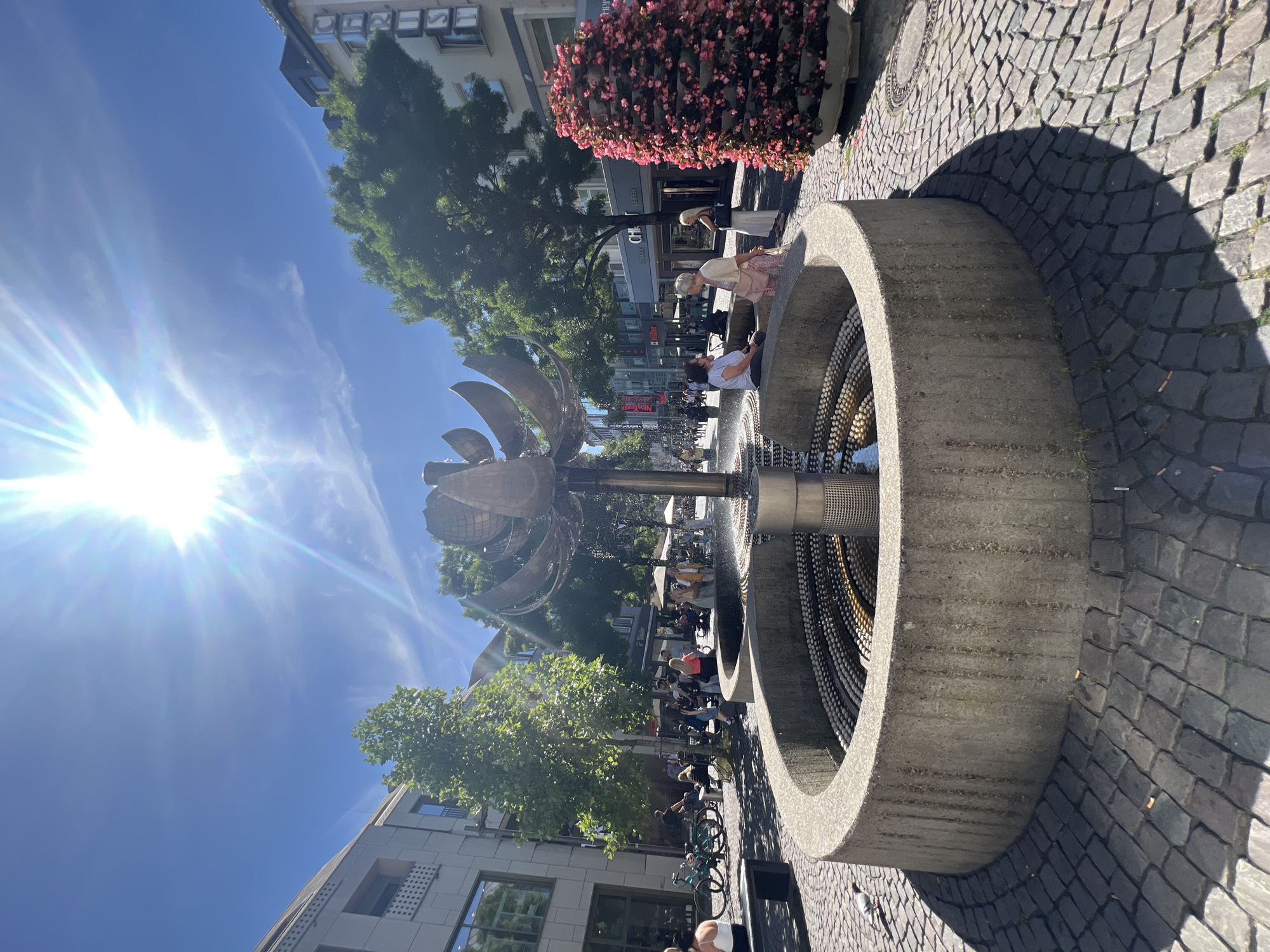 This is an outdoor shot of a fountain in a city, likely Aachen, Germany, on a sunny day. The sun is bright and positioned in the upper left of the frame, casting long shadows across the cobblestone pavement.
The main subject is a circular, concrete fountain with metal elements. Inside the fountain, there are multiple tiers of what appear to be small, round objects, possibly beads or stones, creating a textured surface. A metallic pipe extends upwards from the center of the fountain.
Several people are visible around the fountain and in the background. Some are sitting on the edge of the fountain, while others are standing or walking nearby. One woman, wearing a pink dress, is seen interacting with the fountain. In the background, there are buildings, trees, and shops lining a street. The presence of parked bicycles suggests a public urban space.
The time of day appears to be midday or early afternoon, given the bright sun and the length of the shadows. The weather is clearly sunny and pleasant. The overall mood is relaxed and social, with people enjoying the outdoor space.
There is some visible text on the buildings in the background, though it is mostly illegible due to the distance and image resolution. One sign appears to have the letters "CH" and "EN" visible. Another sign has "WIR" in red letters.