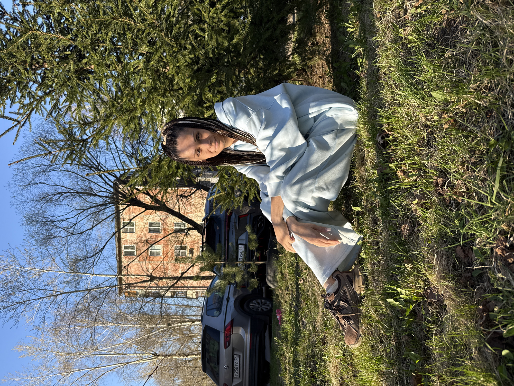 A woman is captured in a squatting or crouching pose on a grassy patch of ground under a clear blue sky. She is positioned slightly to the right of the center in the foreground. She has long, dark braided hair and is wearing a light blue, casual tracksuit consisting of a hooded sweatshirt and sweatpants. Her footwear appears to be dark-colored athletic shoes with light soles. She looks directly at the camera with a neutral to slightly smiling expression.
Behind the woman, a dense evergreen tree, possibly a fir or spruce, dominates the midground, its dark green needles contrasting with the bright sunlight. Further back and to the left, a deciduous tree with bare branches is visible against the clear sky, suggesting early spring or late autumn. In the background, a multi-story building made of light brown or brick material with numerous windows can be seen to the left. Several cars are parked in the background, including a silver SUV with the Russian license plate "Y258YO76" and another darker SUV with a partially visible license plate "0KM44".
The lighting indicates a sunny day, likely late morning or early afternoon, with distinct shadows cast by the trees and the woman herself. The overall setting appears to be an outdoor, possibly urban or suburban, park-like area or residential space in Rusanovo, Russia, as suggested by the car license plates and provided context. The grass is green with some dry patches and sparse weeds.