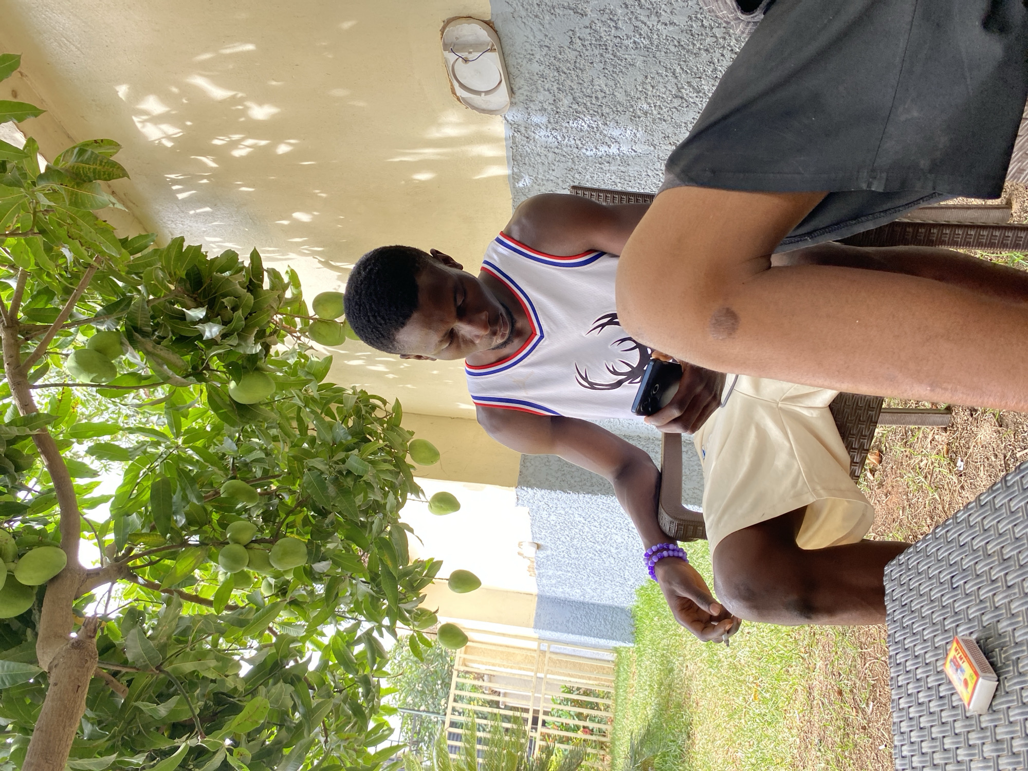 Outside on what appears to be a patio in Barakin Akawo, Nigeria, a dark-skinned man wearing a white tank top with red and blue trim is seated and looking at a mobile phone he is holding in his hands. There is a tree laden with green fruits, possibly mangoes, to his left. Another person, wearing black shorts and light-colored shorts is partially visible. A box of matches is on the table next to him, and he is wearing a purple bracelet. The background shows a yard, greenery, and a building.