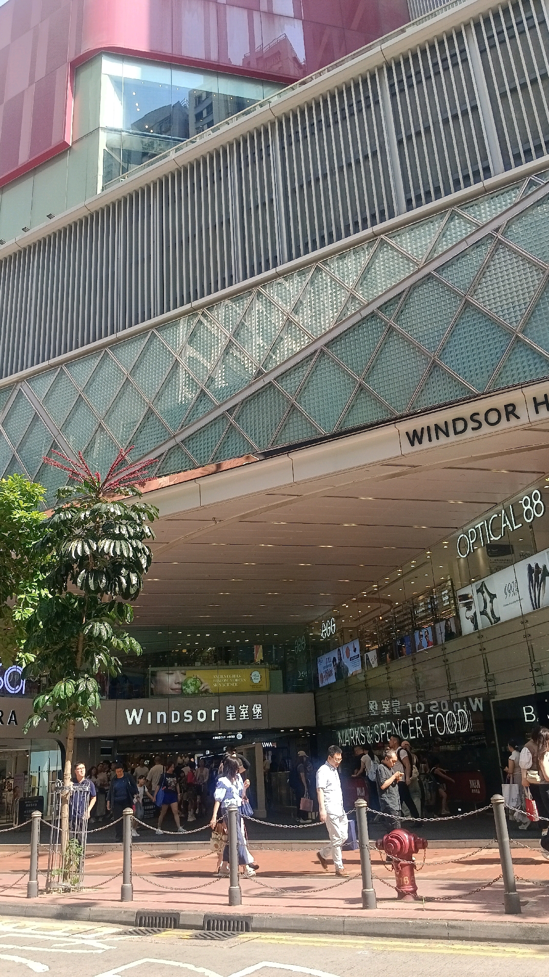 The image depicts the exterior of a modern commercial building, likely a shopping mall or department store, in Hong Kong, China. The building features a facade with horizontal metal louvers, large glass panels, and a distinctive diagonal patterned glass section. A prominent sign reads "WINDSOR" in large black lettering, with Chinese characters beneath it, suggesting a retail establishment. Below this, another sign displays "OPTICAL 88". The ground level shows a busy entrance with many people walking in and out of the building. Various shop signs are visible, including "MARKS & SPENCER FOOD". The scene is set on a sunny day, with a tree and a red fire hydrant visible on the sidewalk. The street level is paved, with a curb and drainage grates. People are dressed in casual summer clothing. The overall impression is a vibrant urban commercial district.