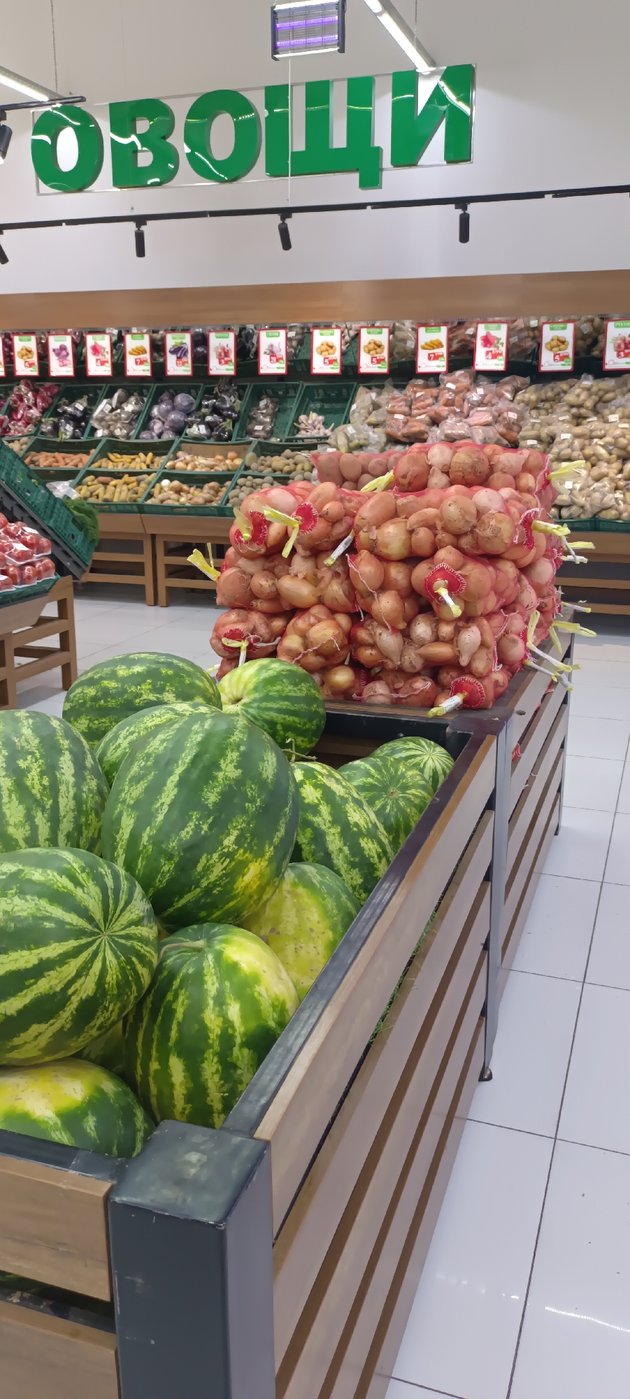 This is a vertical, full shot of a produce section in a supermarket. In the foreground, a wooden bin is filled with several whole watermelons. To the right of the watermelons, a large pile of onions in red mesh bags is displayed on a wooden shelf. In the background, various vegetables like potatoes, carrots, and eggplants are neatly arranged in green crates on shelves. Above the produce, a large green sign with white lettering reads "ОВОЩИ" which is Russian for "Vegetables." The lighting appears to be from overhead fluorescent lights. The floor is tiled in a grid pattern. The scene is static and shows a typical retail environment for fresh produce.