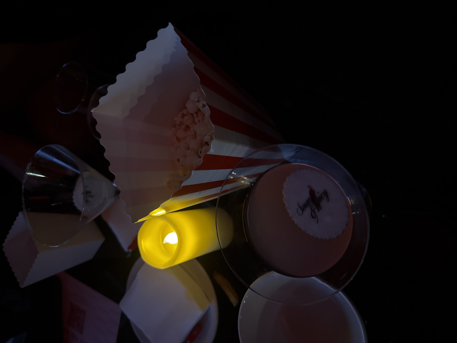 A table is set with illuminated decor. A striped red and white popcorn container sits near a martini glass, adding a cinematic touch. Next to this is a circular cake on a transparent plate, decorated with an edible logo "SugarAffairy". A cylindrical candle is positioned to give a soft, yellow light. The entire scene appears to be reflected on a glossy surface, contributing to the atmosphere.