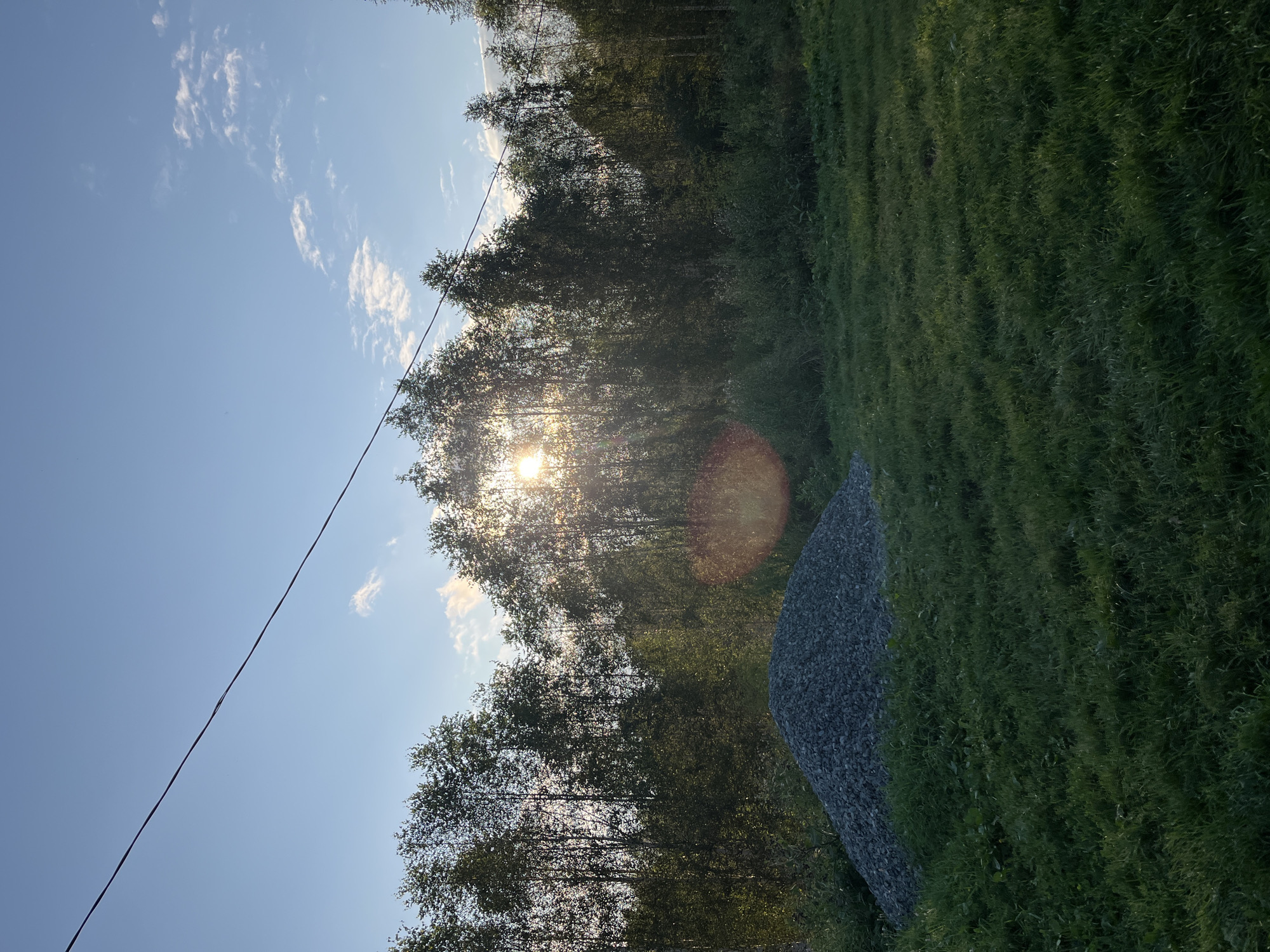 A pile of gravel sits in a grassy field under a clear blue sky with scattered clouds. A line of trees forms a dense backdrop, with the sun shining through them, creating a lens flare effect. A power line stretches across the top of the frame. The scene appears to be in Nizhny Tagil, Russia.