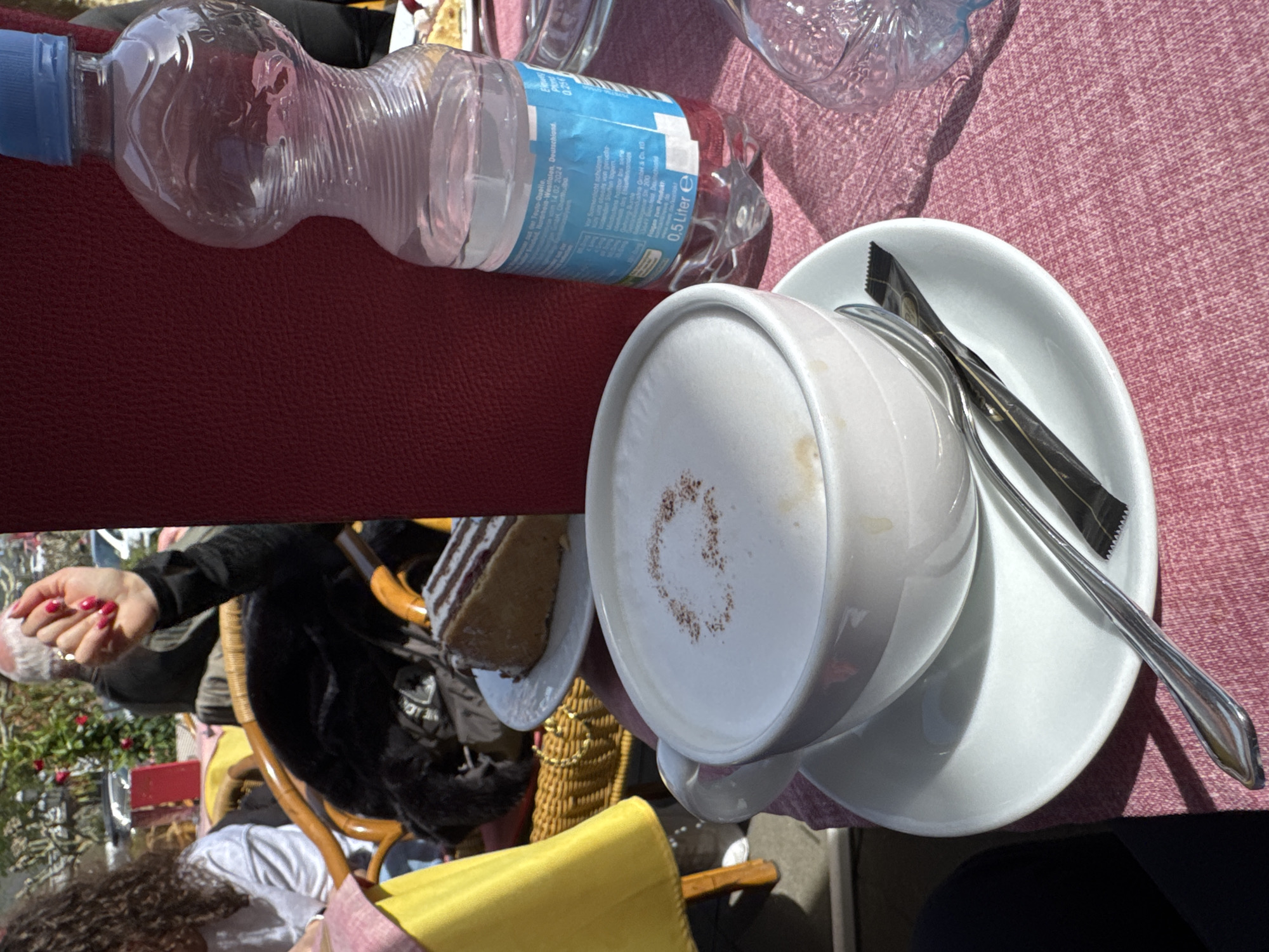 Here's a detailed description of the image:
**Overall Scene:** The image is taken from a high-angle, slightly off-kilter perspective, showing a scene at what appears to be an outdoor café or restaurant.
**Objects:**
* A partially consumed bottle of mineral water (branded "Teuto-Quelle") is prominently positioned at the top left.
* Next to the water bottle, and partially off-frame, is a likely empty, decorative glass water pitcher.
* A cup of coffee (likely a cappuccino) sits on a white saucer, complete with a spoon and a sugar packet on the right side of the frame. The coffee has a latte art pattern of a coffee bean design on the surface.
* A plate with a piece of layer cake is present on the left side.
**People:**
* Parts of at least two individuals are visible. One wears a yellow garment and the other is wearing a black garment with painted nails.
**Environment:**
* The table is covered with a pink tablecloth.
* The setting is outdoors, judging by the presence of chairs, foliage, and dappled sunlight.
* In the background, glimpses of a city scene are present.
**Location (Based on OCR Text):** The brand of water (Teuto-Quelle) indicates the location is likely in Germany, specifically in Cochem, where many of the water brands are produced.