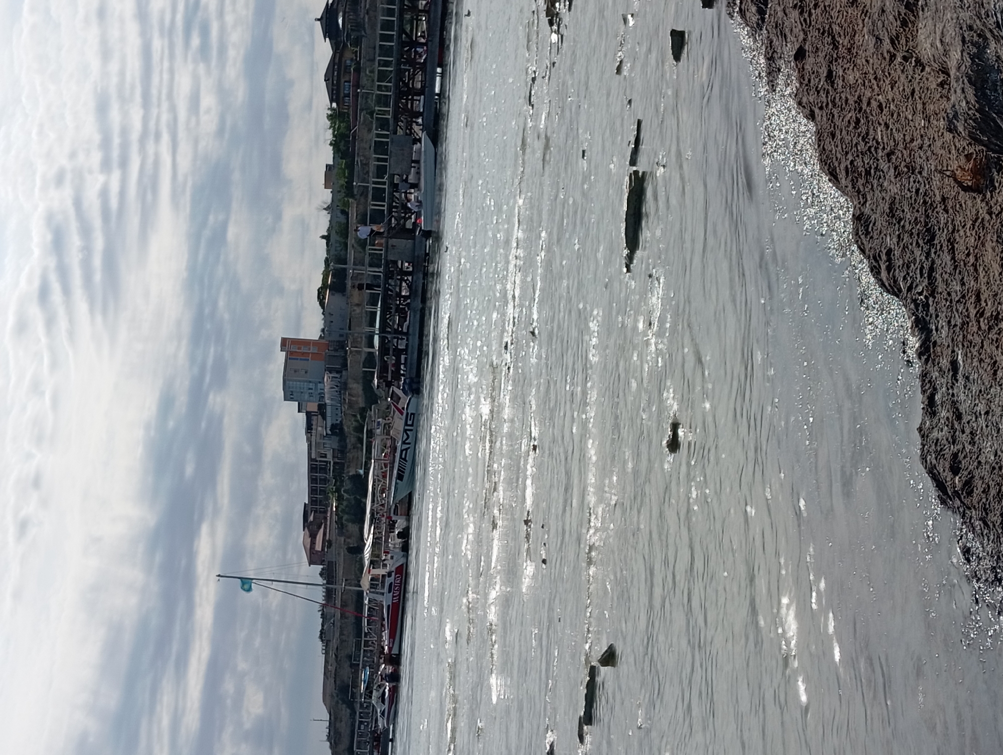 The image depicts a waterfront scene in Aktau, Kazakhstan, likely along the Caspian Sea coast. In the foreground, the water is choppy with reflections of the sky. Several boats are moored at a pier or jetty in the mid-ground. One boat prominently features the word "MAESTRO" and another has "AMG" written on its side. A sailboat with a turquoise and white flag is also visible. In the background, buildings line the shore, some appearing to be residential or hotels, situated on a slightly elevated terrain. The sky is overcast with clouds. The shoreline in the immediate foreground consists of dark, rough material, possibly rocks or artificial breakwaters, with some debris floating in the water. A few people are visible on the pier, and a couple of birds are in the water. The overall atmosphere suggests a moderately busy day at a coastal location.