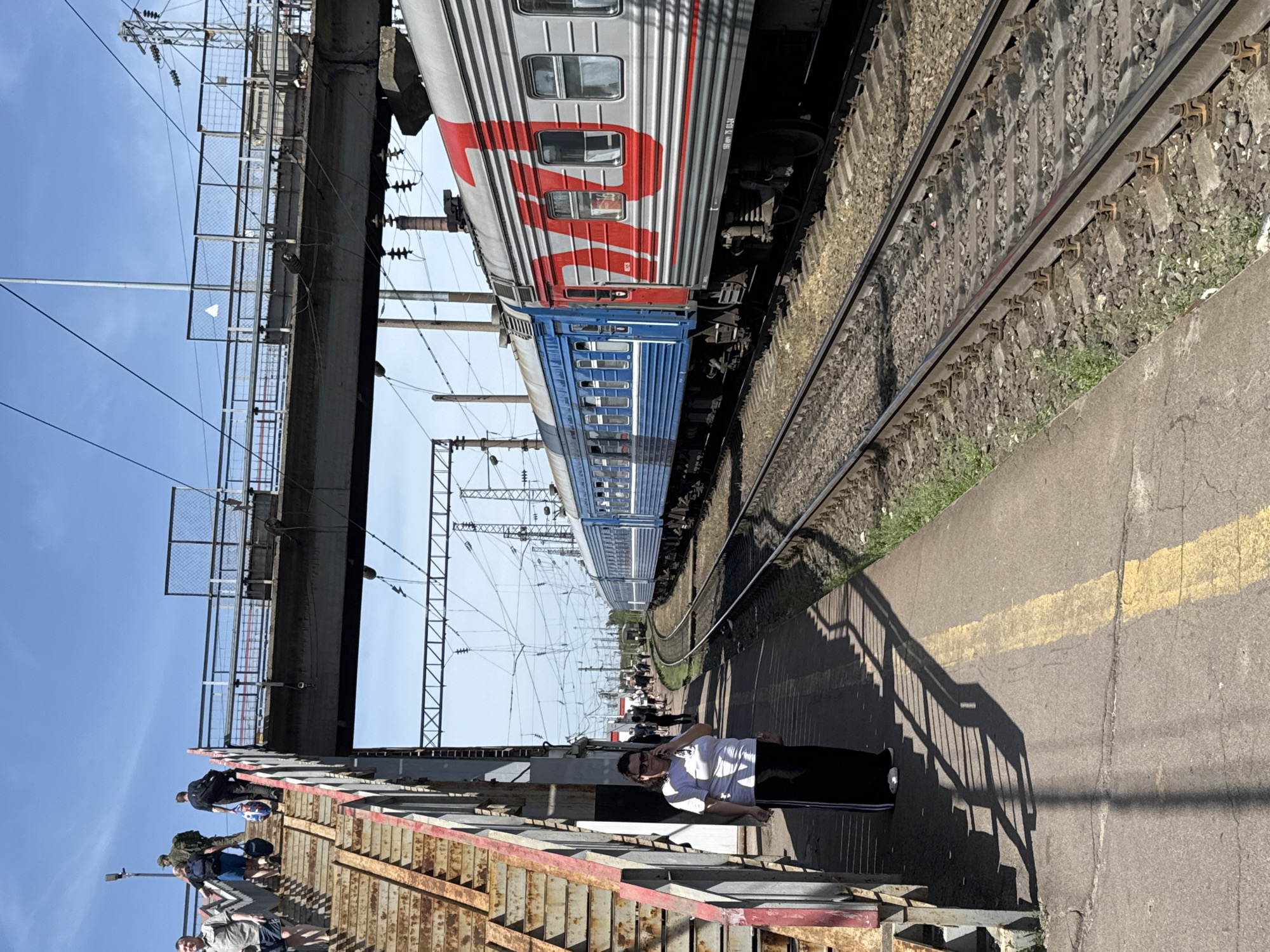 A railway station in Krasnodar, Russia, is visible in the image. An electric train, painted in gray, red, and blue, is running along the tracks parallel to the platform. The railway infrastructure includes overhead electrical lines and a bridge above the tracks. A person stands on the platform, and others are on the elevated walkway. The platform has a yellow safety line marking its edge. The sky is clear and blue.