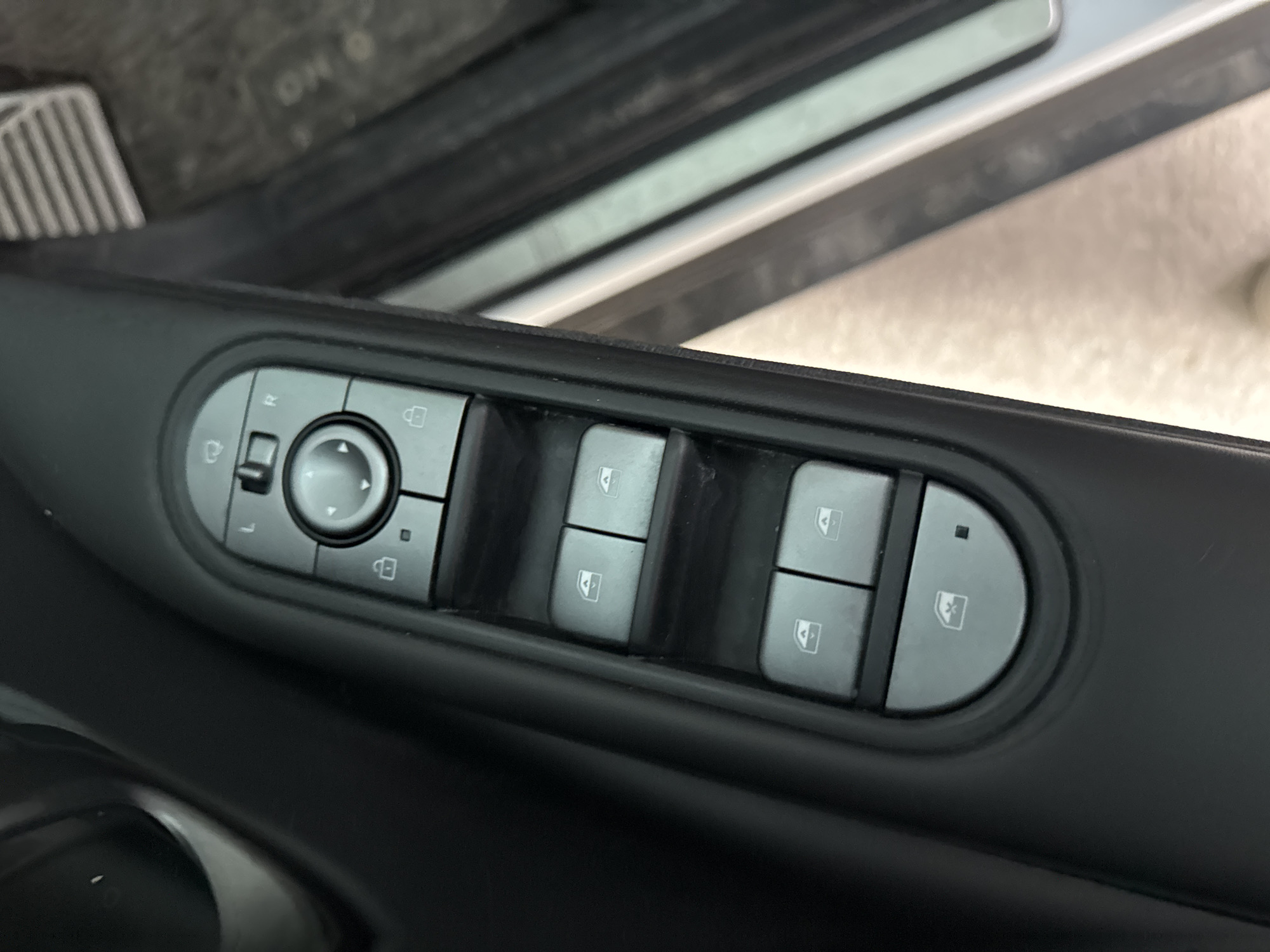 The image shows a close-up of a car's interior, specifically the driver's side door panel with window and mirror controls. The panel is black and houses a series of buttons. At the top, there is a circular control with 'L' and 'R' labels, suggesting it's for mirror adjustment. Below this are buttons with icons of a closed padlock and an open padlock, indicating lock and unlock functions. Further down, there are four rectangular buttons, each with an icon of a car window. The bottom-most button in the window control cluster has an additional icon that appears to be a window with a diagonal line through it, possibly indicating a child lock feature for the windows. The background shows a blurred view of the car's floor mat and the car's door frame. There are no people visible in the image. The lighting suggests an indoor setting, possibly a garage or the interior of a parked car. The image is taken from a slightly elevated angle, looking down at the controls. This image does not provide any information about the city of Shumen, Bulgaria.