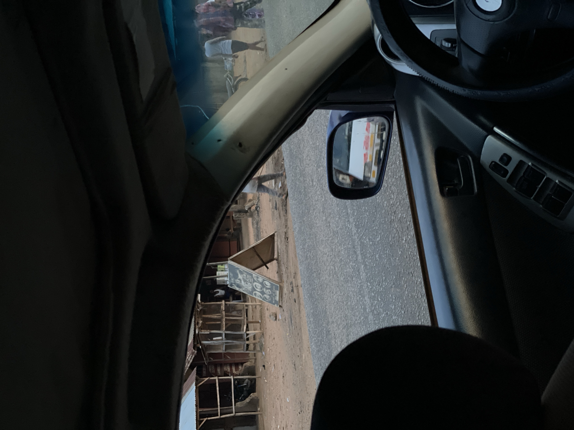 View from inside a car in Akwanga, Nigeria. The steering wheel and part of the dashboard are visible. The side mirror reflects a building. Outside the window, there's a dusty road. A man is seen walking with a bicycle and another person is visible further away. There's a building with signs, possibly a local business like a barber shop. The sky is visible through the windshield.