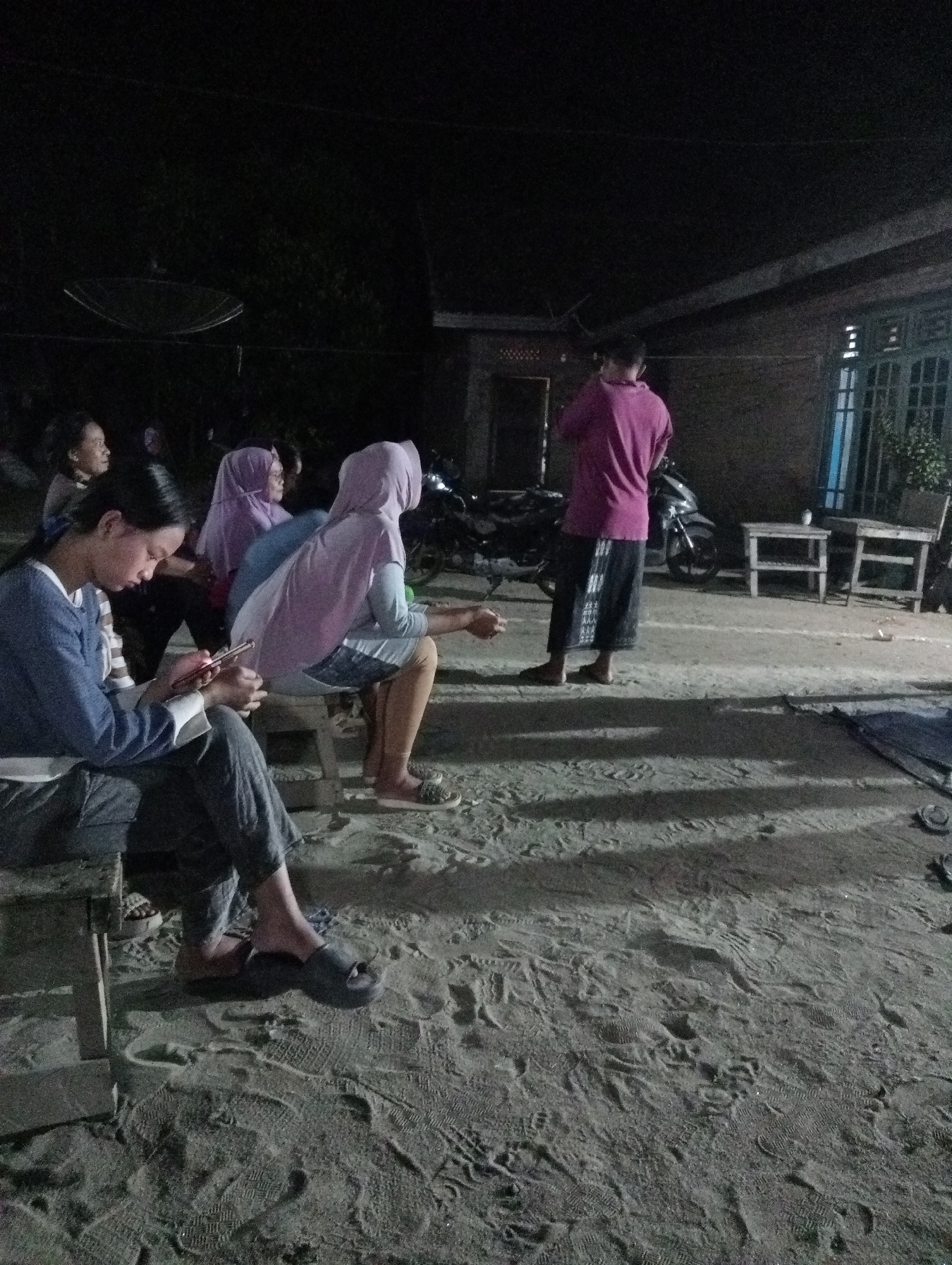 In the dimly lit evening, several individuals are gathered outdoors in Rasuan Baru, Indonesia. In the foreground, a young woman wearing a blue top and dark pants is seated on a wooden bench, engrossed in her mobile phone. Beside her, another person in a light blue hijab and a pink outer garment is also seated. Behind them, two other individuals, one in a light purple hijab and another with a darker hijab, are visible. To the right of the group, a person wearing a magenta shirt and a sarong stands near a motorcycle and two small wooden tables. The ground is sandy and covered with numerous footprints, indicating recent activity. In the background, a satellite dish is mounted on a pole, and a residential building with a distinctive window design is present. The overall atmosphere suggests a casual gathering or waiting period in an outdoor setting.