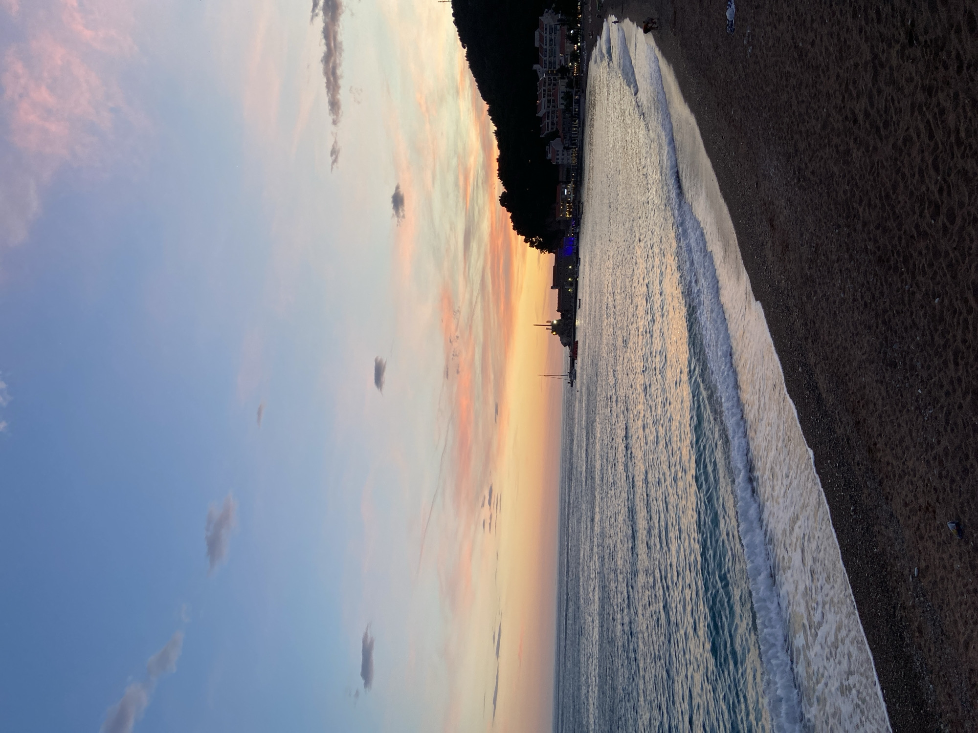 A vertical-format shot shows the beach at Petrovac centar, Montenegro, at sunset. To the left is a gradient sky: light blue transitioning to peach and orange hues near the horizon. Scattered clouds appear in the sky, some colored with shades of pink. The sea extends from the lower right corner to the center, reflecting the colors of the sunset. The water's surface has gentle waves. The beach, composed of dark sand and pebbles, occupies the right side of the image. Along the shoreline, the foamy edges of the waves are visible. In the middle, the silhouette of a hill covered in trees is seen, with buildings and lights along the coastline below. A sailboat is visible in the sea. People are barely visible along the shore.