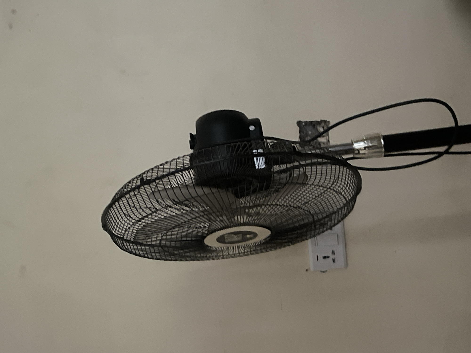 The image captures a close-up view of a black wall-mounted or ceiling-mounted oscillating fan against a light, neutral-colored wall. The fan is the primary subject, positioned towards the lower left and angled upwards. It features a black metal mesh cage and a black motor housing. In the center of the fan's front grille, there's a circular cap, but any potential logo or text on it is not discernible.
The fan is supported by a metallic arm extending from its motor housing to what appears to be a mounting point on the wall or ceiling structure. A black electrical cable is visible, emerging from the fan's assembly and looping loosely before extending further out of the frame to the right.
On the right side of the frame, mounted on the wall, is a white electrical outlet. This outlet appears to be a multi-standard type, designed to accommodate different plug configurations, with visible receptacles that resemble both Type C/F (European) and Type B (North American) styles. There are faint smudges or marks visible on the light-colored wall, particularly in the upper left corner.
No people are present in the image, and no specific activity is taking place; the fan is not visibly operating. The time of day, weather, and specific emotions are not discernible from the photograph. No explicit text is clearly legible on any of the objects within the image. Given the location context of Howayi, Nigeria, the multi-standard electrical outlet is a notable detail, as such outlets are commonly used in various regions, including parts of Africa, to provide versatility for different appliance plug types.