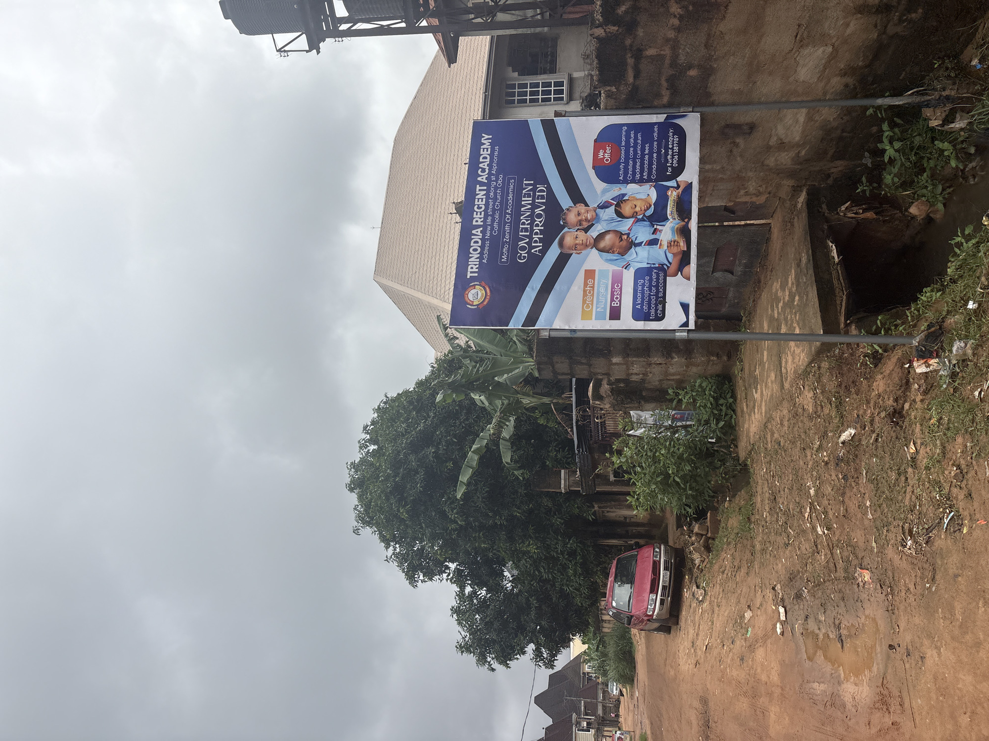 The image depicts a street scene in Obosi, Nigeria, under an overcast sky. A red vehicle is parked on the left side of a dirt road, which appears to be in poor condition with puddles and scattered debris. To the right of the road, a large billboard advertises "TRINODIA REGENT ACADEMY," listing its address as "New life street along st Alphonsus Catholic Church Oba" and its motto as "Zenith Of Academics." The billboard also highlights that the school is "GOVERNMENT APPROVED!" and details the offered programs: Crèche, Nursery, and Basic. It further lists the school's offerings such as "Activity based learning," "Christian core values," "Updated curriculum," and "Affordable fees." Contact information for further inquiry is also provided on the billboard. Behind the billboard and the street, a building with a distinctive roof structure is visible, along with lush green trees and vegetation. A large black water tank on stilts stands to the far right. The overall environment suggests a developing urban or semi-urban area.