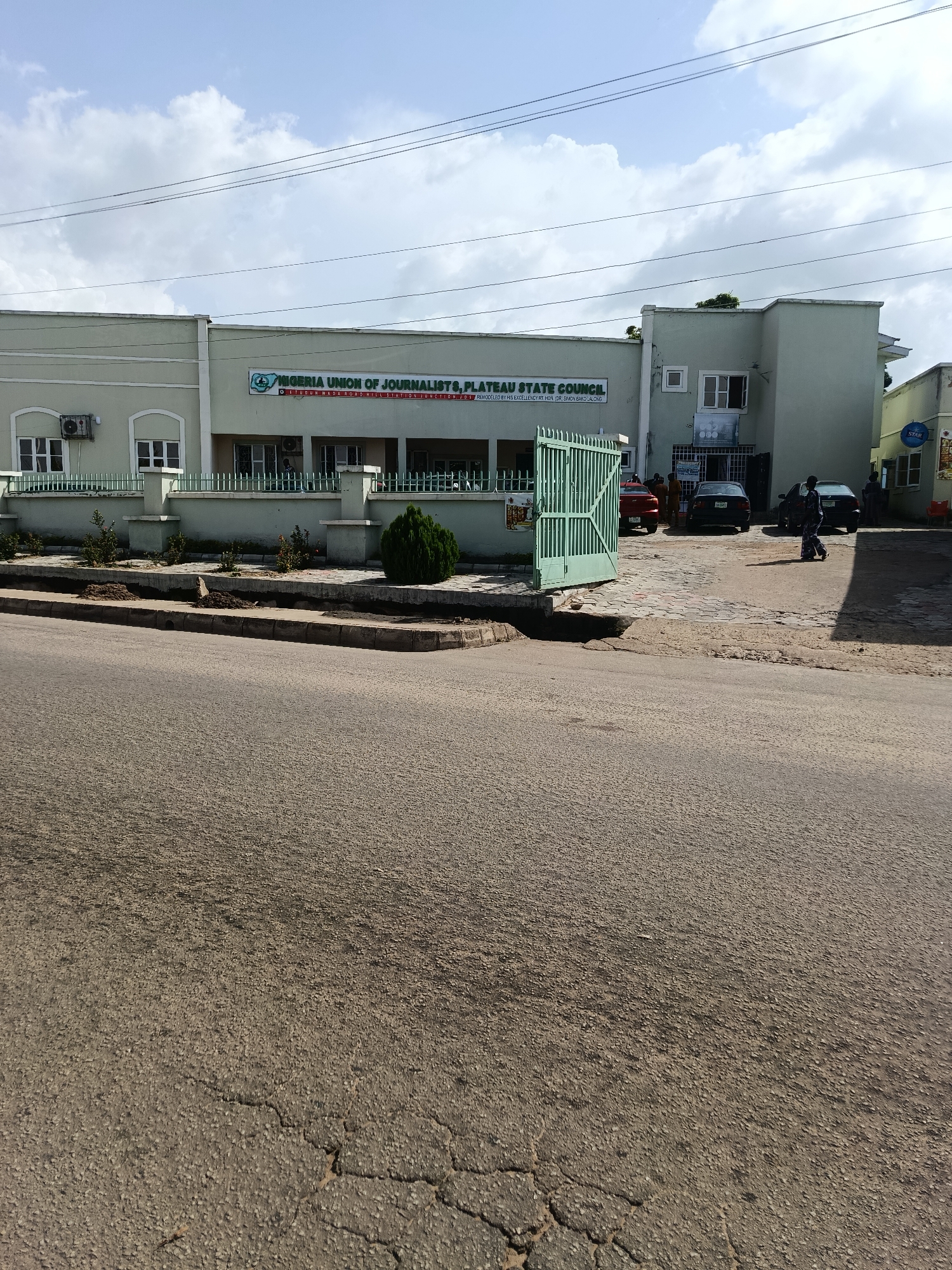 This is a full-shot, eye-level, outdoor photograph of the Nigeria Union of Journalists, Plateau State Council building, located at Old Airport Road, Hill Station Junction, Jos, Nigeria. The building is a light green, two-story structure with a sign displaying "NIGERIA UNION OF JOURNALISTS, PLATEAU STATE COUNCIL" and the text "REMODELED BY HIS EXCELLENCY RT. HON. DR. SIMON BAKO LALONG." In front of the building is an open, green metal gate and a paved courtyard with several parked cars, including a red sedan, a dark sedan, and a black sedan. Three individuals are visible in the courtyard: two standing near the cars and one person walking away from the camera towards the street. The building has several windows and air conditioning units visible on its facade. The foreground of the image shows a cracked asphalt road and sidewalk. The sky is partly cloudy, with distinct white clouds against a blue background. The scene suggests a daytime setting, likely during daylight hours due to the clear lighting.