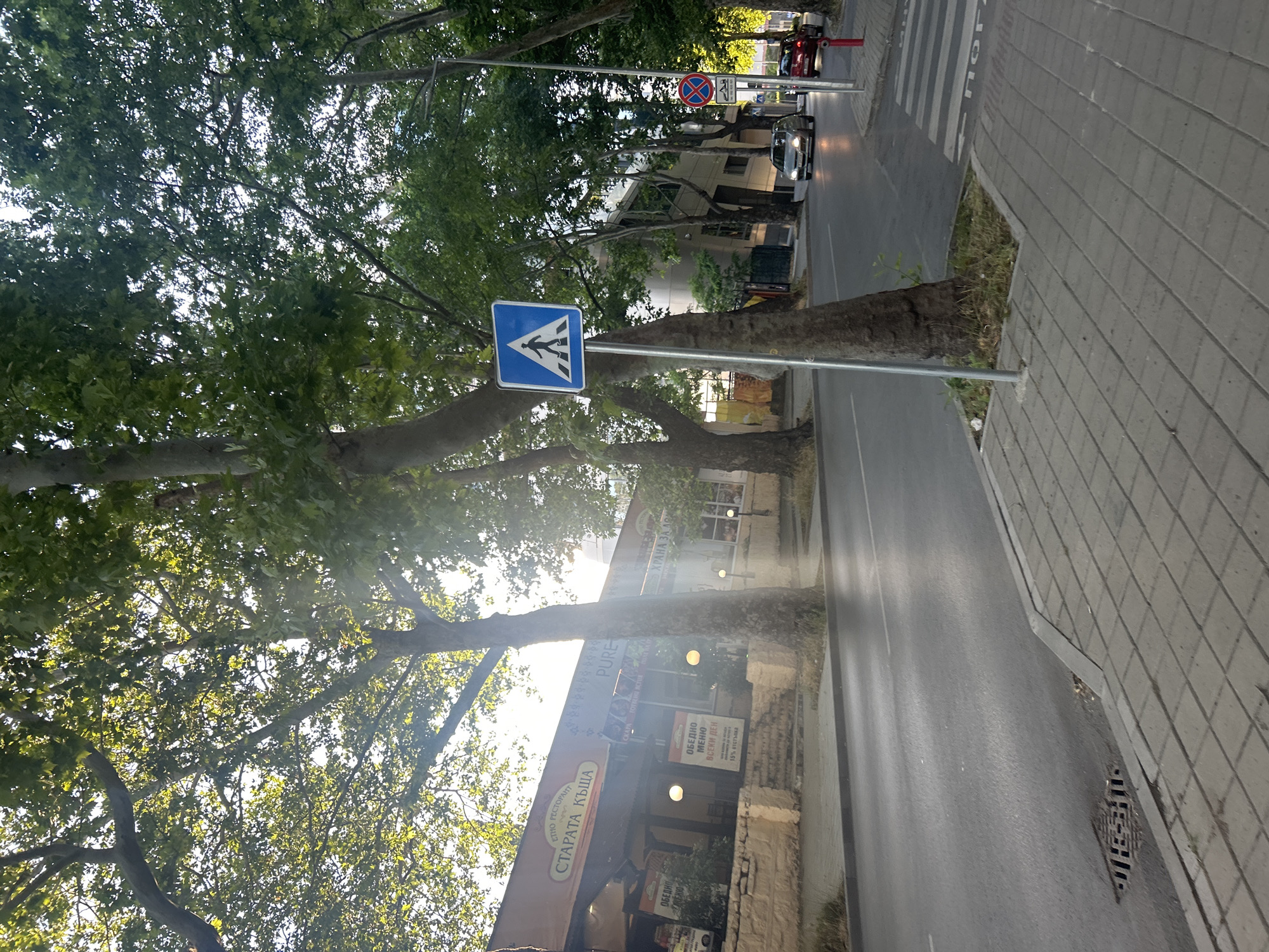 An eye-level, medium shot captures a street scene in an urban environment, likely in the late afternoon due to the angled sunlight filtering through a dense canopy of mature trees. The foreground is dominated by a paved sidewalk composed of light gray square tiles. To the right of the sidewalk, a road stretches forward, marked with a white pedestrian crossing. A prominent blue and white pedestrian crossing sign stands on a metal pole on the sidewalk, indicating a designated area for people to cross the street.
The road is asphalt and appears wet in places, reflecting the sunlight. Several cars are visible on the road, with one silver car in the mid-ground and a red car further back. A red fire hydrant is positioned near the right edge of the frame.
Along the left side of the street, a building with an orange awning stretches across the frame. Signs on the building indicate it is a restaurant named "СТАРАТА КЪЩА" (The Old House). The building's facade is decorated with various posters and images. The architecture appears to be a mix of traditional and modern elements, with a stone base supporting the main structure.
The overall impression is one of a well-maintained urban street with a pedestrian-friendly environment, shaded by abundant greenery. The lighting suggests a pleasant, possibly warm, day.