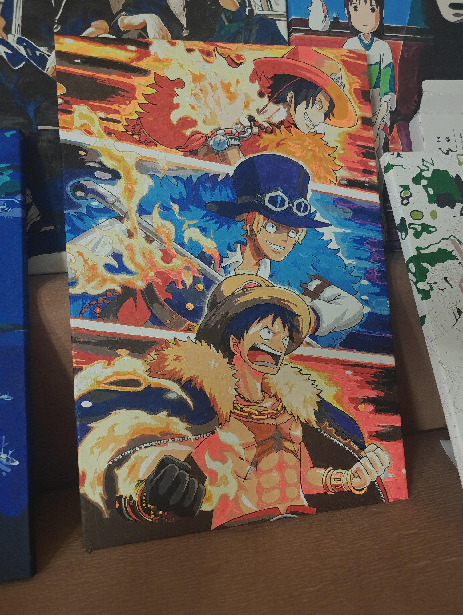 The image depicts a painting of three anime characters arranged vertically. The topmost character wears a red hat and has spiky black hair, surrounded by red and orange flames. The middle character has blonde hair, wears a blue hat with goggles, and is surrounded by blue flames. The bottom character has a straw hat, bare chest, and is also surrounded by orange flames. The background consists of horizontal red and blue stripes. The painting is resting on a brown surface, possibly a table or shelf. Other paintings are visible in the periphery. The location is an indoor setting.