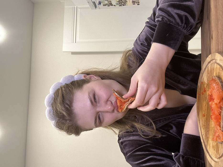 A woman with a blue padded headband is eating a piece of bread topped with red caviar. She wears a dark velvet robe or jacket. A wooden cutting board with more bread and caviar is visible to the right. The background includes a pale wall and a white cabinet. The image appears to be taken indoors with bright lighting.