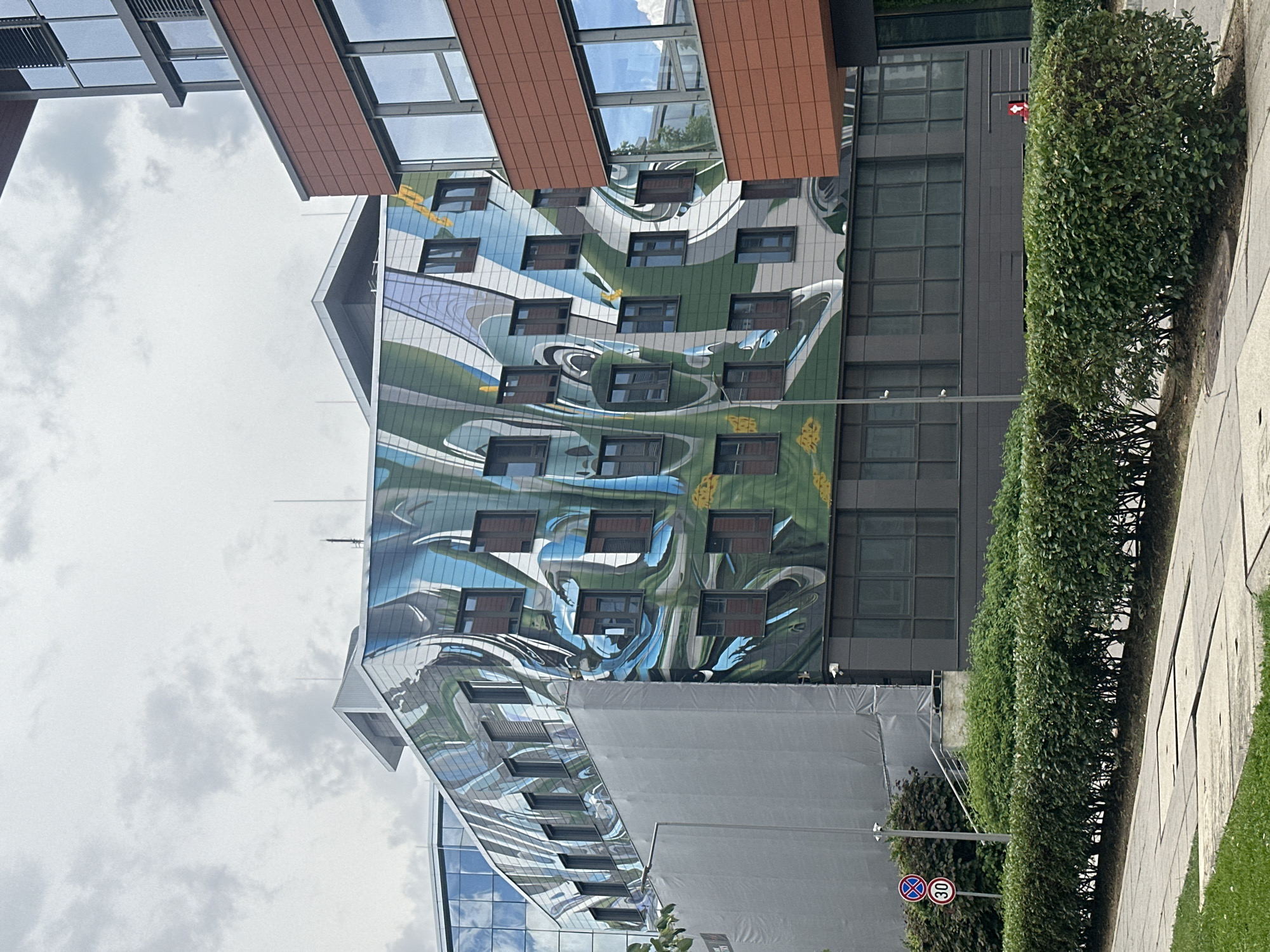 The image shows a modern office building in Sofia, Bulgaria, featuring a striking mural on its facade. The mural is an abstract design in shades of green, blue, and white, with wavy lines and organic shapes that create a dynamic and fluid appearance. The building itself has a contemporary architectural style with large windows and a metallic-looking exterior. To the right of the muraled building, a section of another building is visible, clad in reddish-brown tiles. In the foreground, a neatly trimmed green hedge runs along a paved walkway. A traffic sign indicating a speed limit of 30 km/h is visible on the left side of the image, near the hedge. The sky above is overcast with clouds.