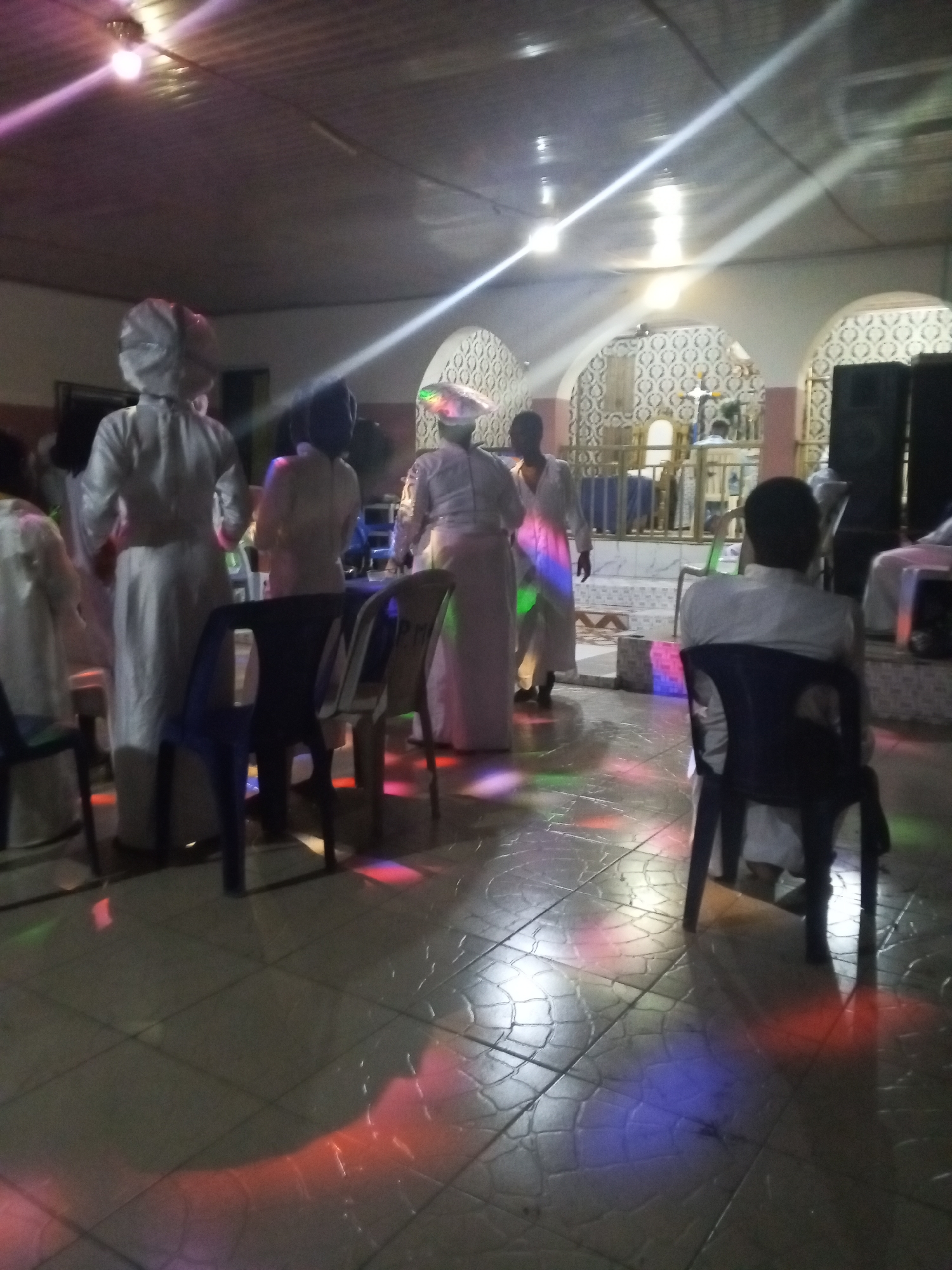 A brightly lit interior space with a tiled floor, reflecting multicolored lights. Several people wearing white robes and head coverings stand and move about in the middle of the room. A few black plastic chairs are scattered around, some with people sitting on them. In the background, there's a raised platform with a decorated railing and a chair, likely an altar or stage. Speakers are visible on the right side.