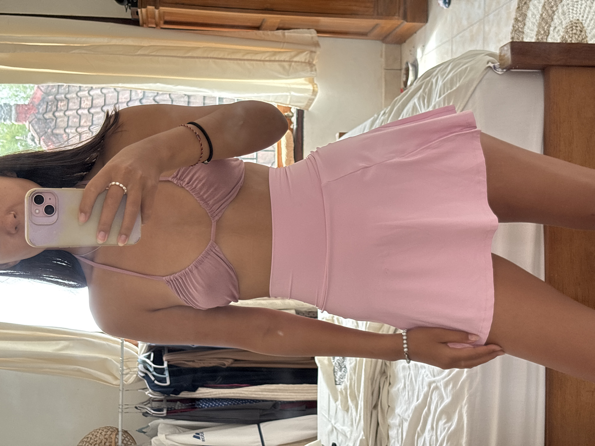 A person is taking a selfie indoors, holding a smartphone up with their left hand. They are wearing a light pink two-piece outfit consisting of a gathered triangle bikini top and a short skirt. The person has dark hair. There is a window behind them, showing what appears to be the exterior of a building with a tiled roof, suggesting a location in Kuta Selatan, Indonesia. Background details also include a wooden dresser, folded clothing on shelves, and bedding with white sheets. The person is positioned in front of a mirror.