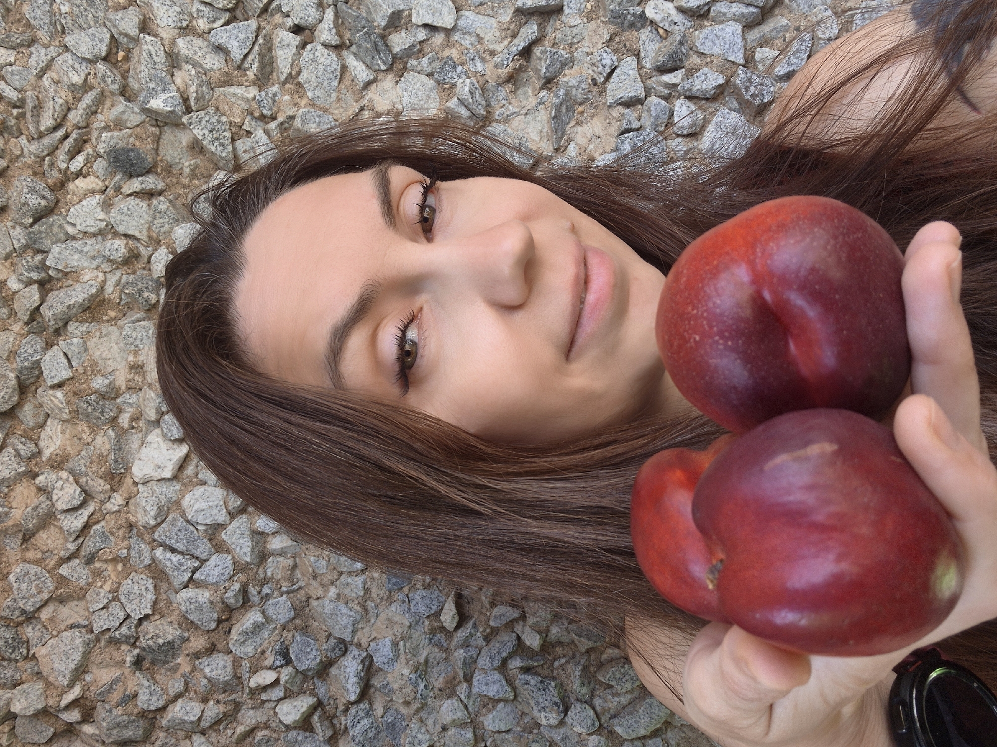 A woman with long brown hair is holding three red nectarines in her left hand, extending them towards the camera. She is looking towards the camera with a slight smile. The background is a rough, textured stone wall. The image appears to be a selfie taken outdoors, as indicated by the natural light and outdoor setting. The woman's attire is not fully visible, but her shoulder and upper chest are exposed. The specific location in Chaikovsky, Russia is not discernible from the visual information provided in the image.