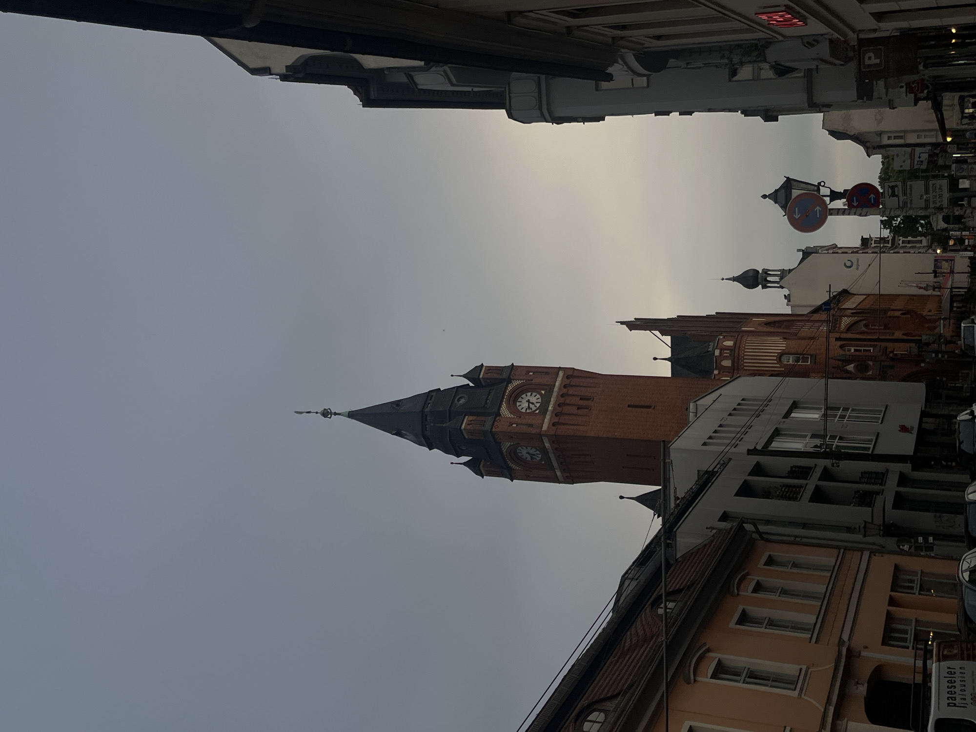 The image captures a street scene in Berlin, Germany, with a prominent brick tower, likely a town hall or church spire, dominating the center of the frame. The tower features visible clock faces and is topped with a dark, conical roof and a spire. Surrounding the tower are various buildings of different architectural styles and colors. To the left, an older building with an orange facade and arched windows is visible, partially obscured by a darker structure. To the right, a taller, more modern building with a gray facade and multiple windows stands. Further down the street, other buildings are visible, including one with a distinctive curved window. Street signs, including traffic signs with red and blue circles and arrows, are present, suggesting a regulated area. The sky is overcast, contributing to a somewhat dim and diffused lighting. The perspective is from street level, looking up towards the tower and buildings. There are no discernible people or animals in the image.