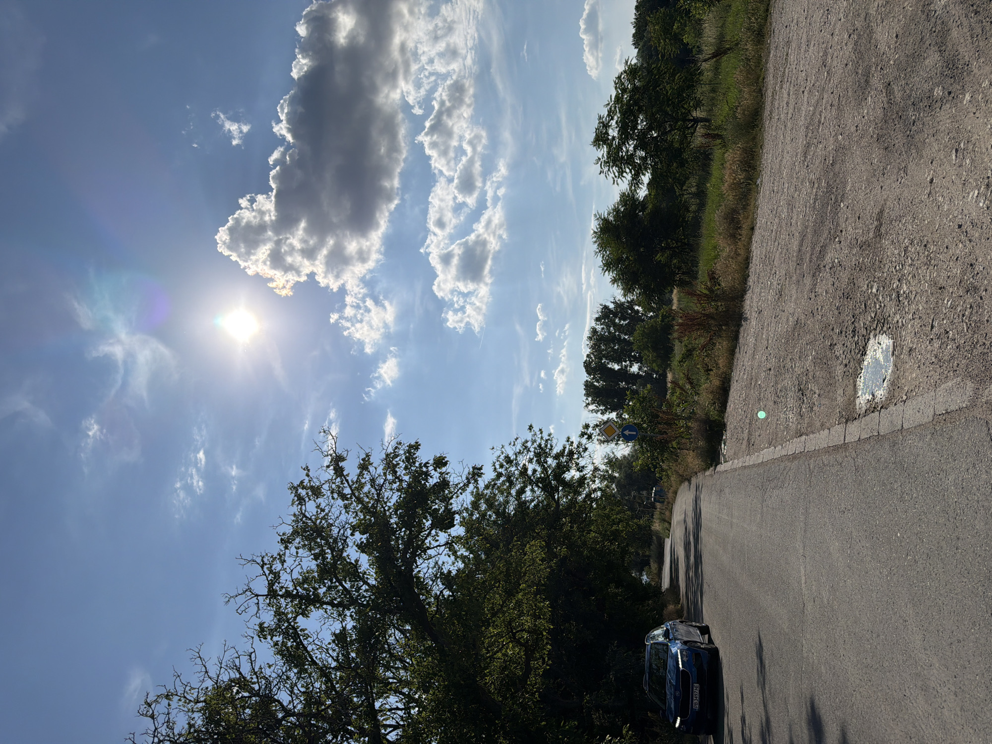 The image depicts a rural road in the vicinity of Plovdiv, Bulgaria. The road surface is asphalt and shows signs of wear, with some potholes visible, particularly on the right side where it transitions to a gravel shoulder. Flanking the road are trees and vegetation, with a prominent large tree on the left casting shadows onto the road. A blue car is parked on the left side of the road. In the distance, more trees and greenery are visible. The sky is bright blue with scattered white and grey clouds. The sun is high in the sky, with lens flare visible, indicating a clear, sunny day. Road signs are present on the right side of the road, including a yellow diamond-shaped sign with a black border and a black arrow pointing upwards, and a blue circular sign with a white arrow pointing upwards. The overall scene suggests a rural or semi-rural setting outside the city center of Plovdiv.