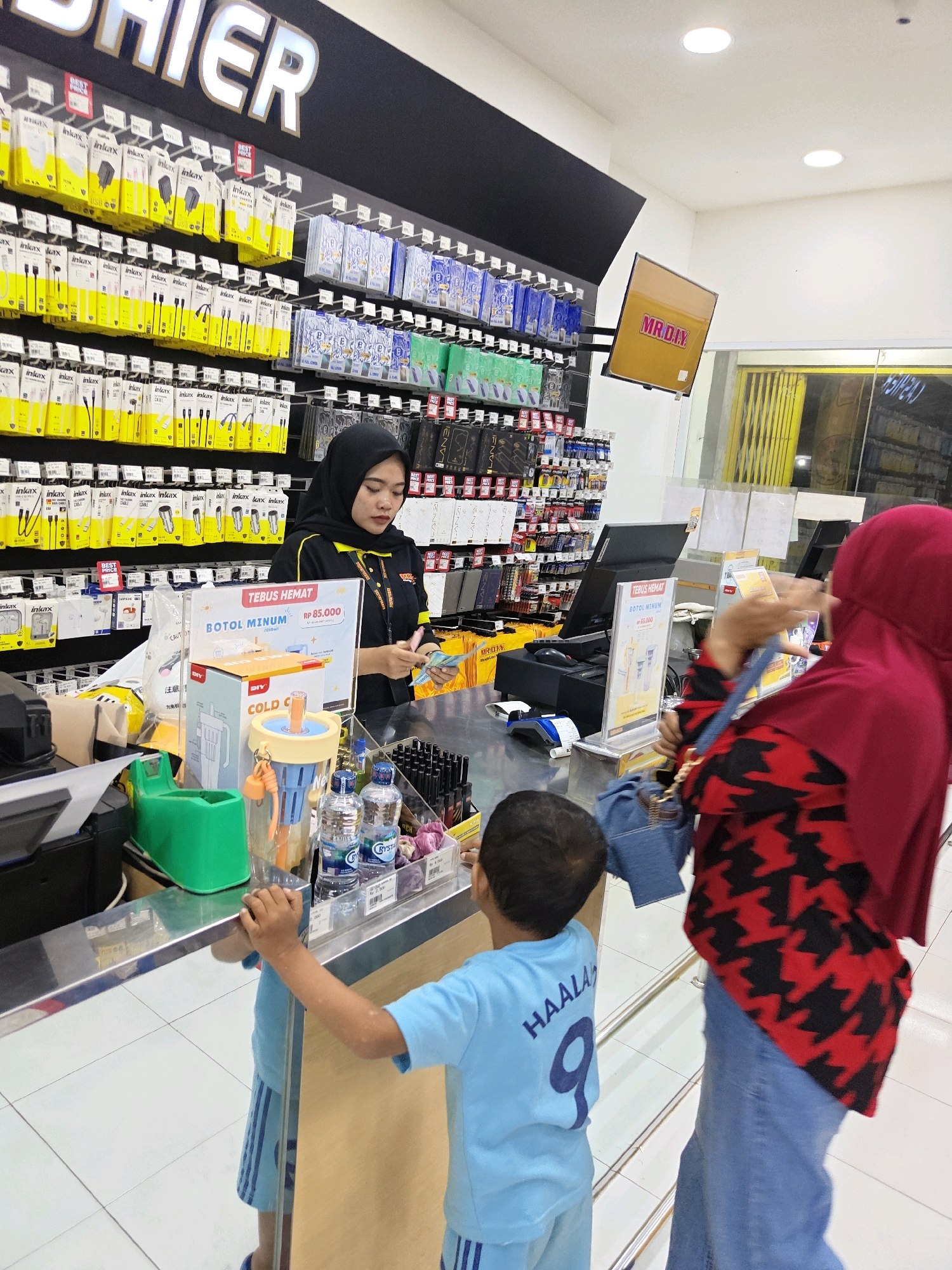 This image depicts the interior of a MR.DIY retail store, likely located in Situbondo, Indonesia. The store specializes in home improvement and hardware supplies, evidenced by the extensive shelving stocked with various products, predominantly yellow packaging with branding such as "inkax." In the foreground, a cashier wearing a black hijab and a black and yellow uniform is processing a transaction at the checkout counter. She is holding cash and appears to be counting it. In front of the cashier is a display for "BOTOL MINUM COLD C," a drink bottle. Two customers are visible: a child in a light blue t-shirt with "HAALAN 9" on the back, standing on the left side of the counter, and a woman wearing a red hijab and a red and black patterned top, standing on the right side, interacting with the cashier. The checkout area includes a point-of-sale terminal and various items for sale, such as bottled water. A television screen mounted on the wall displays the "MR.DIY" logo. The environment is brightly lit, characteristic of a retail setting.