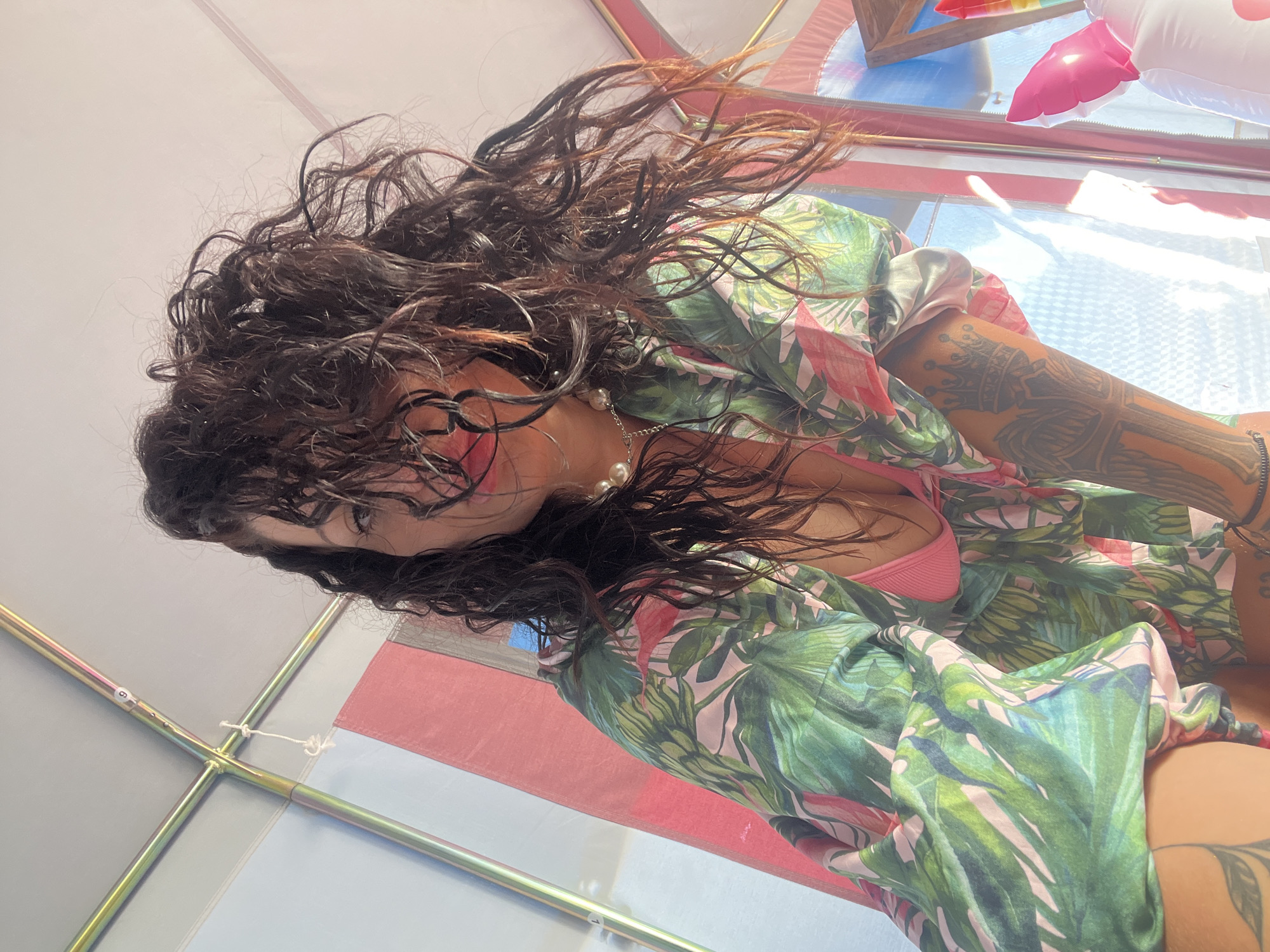 A woman with long, dark, wet curly hair, wearing a pink bikini and a tropical print shirt, is photographed from a low angle inside a tent. Her hair partially obscures her face, but her eyes and lips are visible. She is wearing a pearl necklace. Her right arm, which is tattooed with a crown and wings, is visible. In the background, there is a striped tent fabric, an inflatable unicorn, and natural light filtering through. The scene appears to be set in a recreational area, possibly near the beach, in Sozopol, Bulgaria.