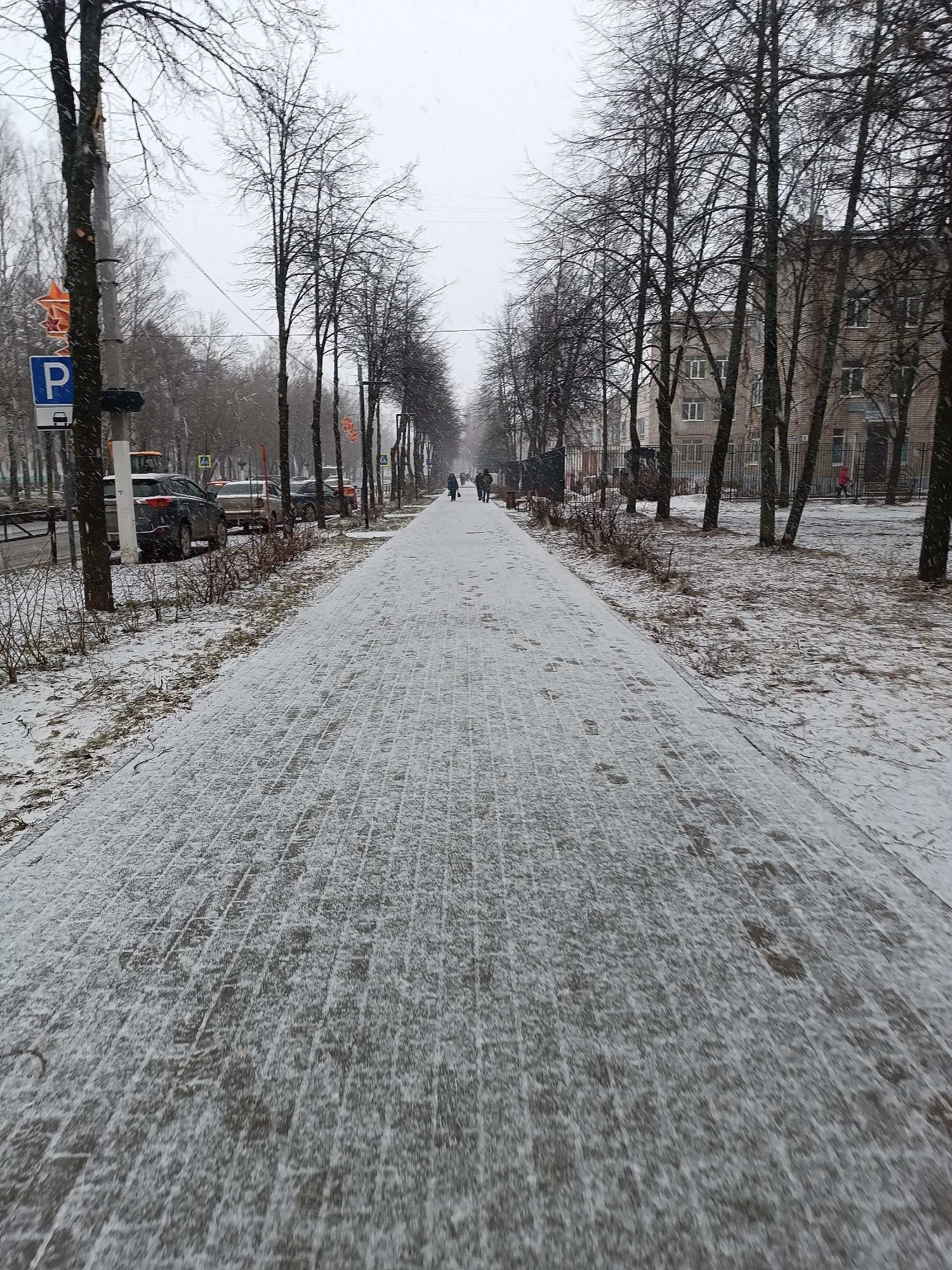 The image depicts a snow-covered brick walkway in an urban setting. The walkway stretches into the distance, flanked by bare trees on either side. Several people are walking away in the distance. Parked cars line the left side of the street, indicated by a "P" parking sign. Buildings are visible behind the trees on the right. The environment suggests winter conditions, with snow covering the ground and walkway. The image appears to be taken at ground level, focusing on the walkway leading into the distance.