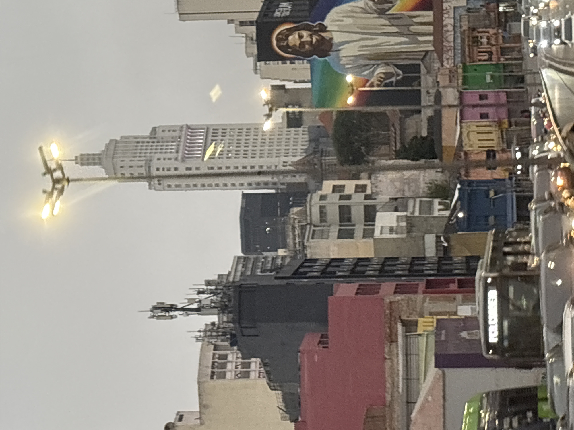 This is a street-level view in São Paulo, Brazil, likely captured from a moving vehicle. The scene is dominated by a dense urban environment with a mix of architectural styles. In the background, a prominent Art Deco skyscraper, the Edifício Altino Arantes (also known as the Banespa Building), stands tall. Adjacent to it are other multi-story buildings, some featuring cell towers on their rooftops.
In the middle ground, a large billboard displays an image of Jesus Christ with outstretched arms, set against a rainbow background. Below this, various colorful buildings line the street, featuring storefronts and multiple windows. A prominent red building is visible on the left.
In the foreground, a bus with illuminated text displaying "POR CURTA" (meaning "short" or "brief" in Portuguese) is partially visible. Several cars are also present on the road, with their headlights and taillights illuminated, suggesting it may be late afternoon or evening. The overall atmosphere is overcast, typical of São Paulo's urban weather.