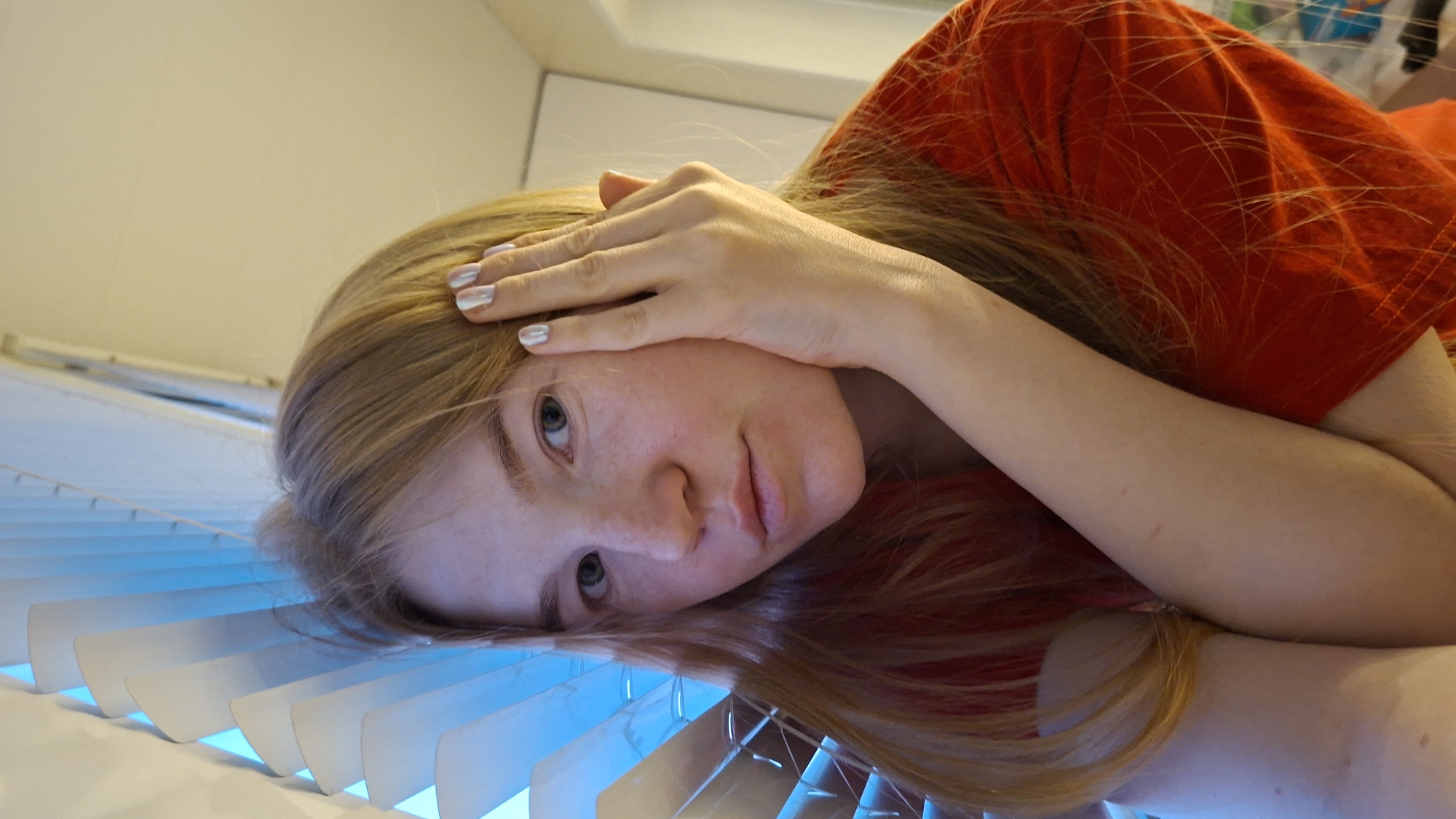 A fair-skinned, red-haired, young woman wearing a red T-shirt lies in an unusual position with her head resting on white venetian blinds. Her right hand is on her forehead. The blinds are illuminated with a blue light. The location of this image is Chaikovsky, Russia.
