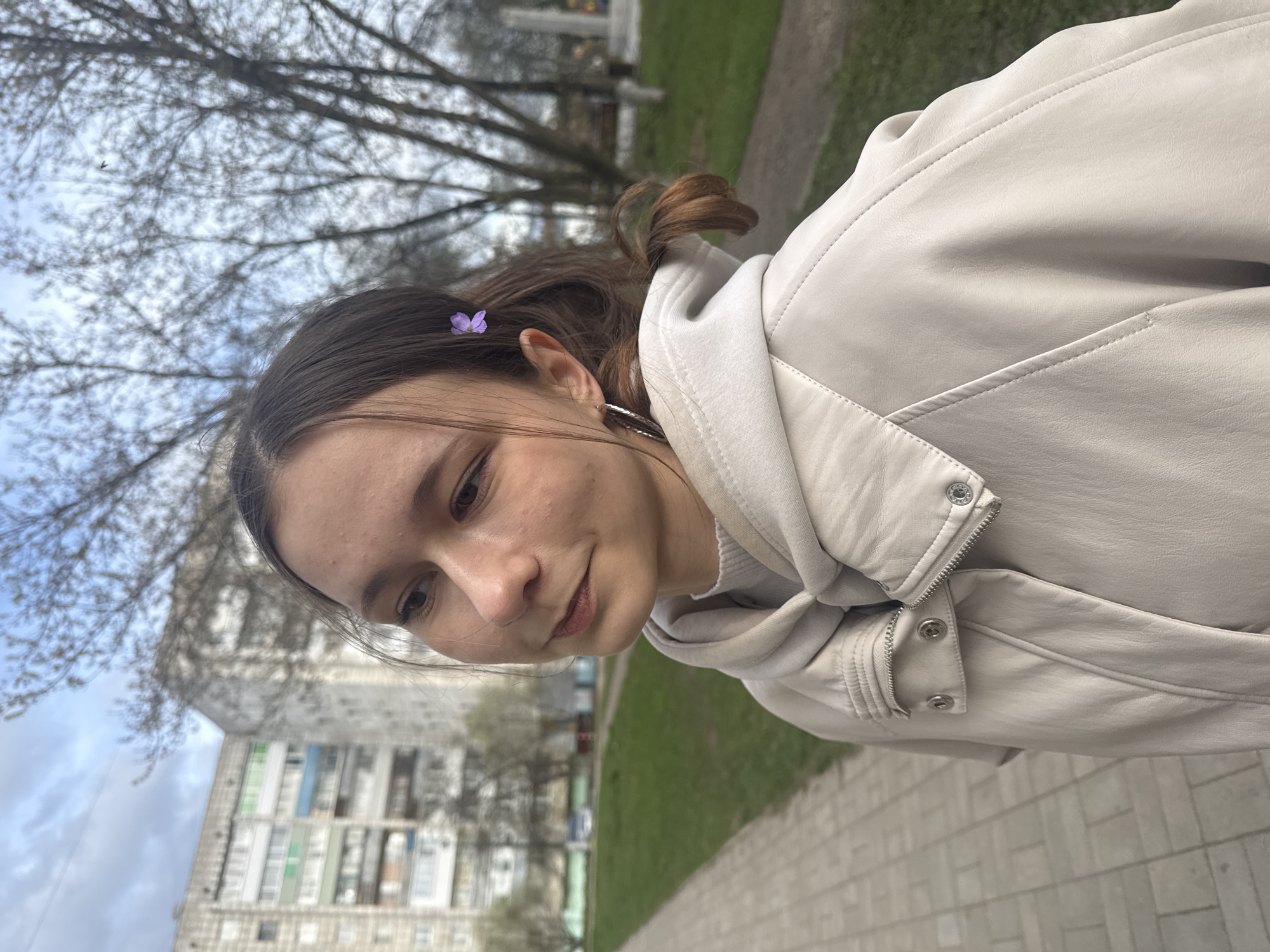 A young woman with brown hair, adorned with a small purple flower near her left temple, is the primary subject of this outdoor portrait. She is wearing a light cream or off-white faux leather jacket over what appears to be a matching hooded sweatshirt. The jacket features zippers, snap buttons, and prominent stitch detailing. Her expression is gentle and soft, with a slight smile as she looks downwards and to her left. Her skin shows visible freckles.
The setting is an urban or suburban environment, consistent with Chaikovsky, Russia. The background features numerous bare tree branches against an overcast sky, suggesting late winter or early spring. In the middle ground, a large, multi-story apartment building with visible colored panels on its facade is partially visible and slightly out of focus. A paved sidewalk or path runs through the scene, adjacent to patches of green grass.
The woman appears to be posing casually for the camera. The time of day is daytime, with soft, diffuse lighting indicating an overcast sky. The image is rotated clockwise, giving a tilted, dynamic perspective to the composition. There is no visible text in the image.