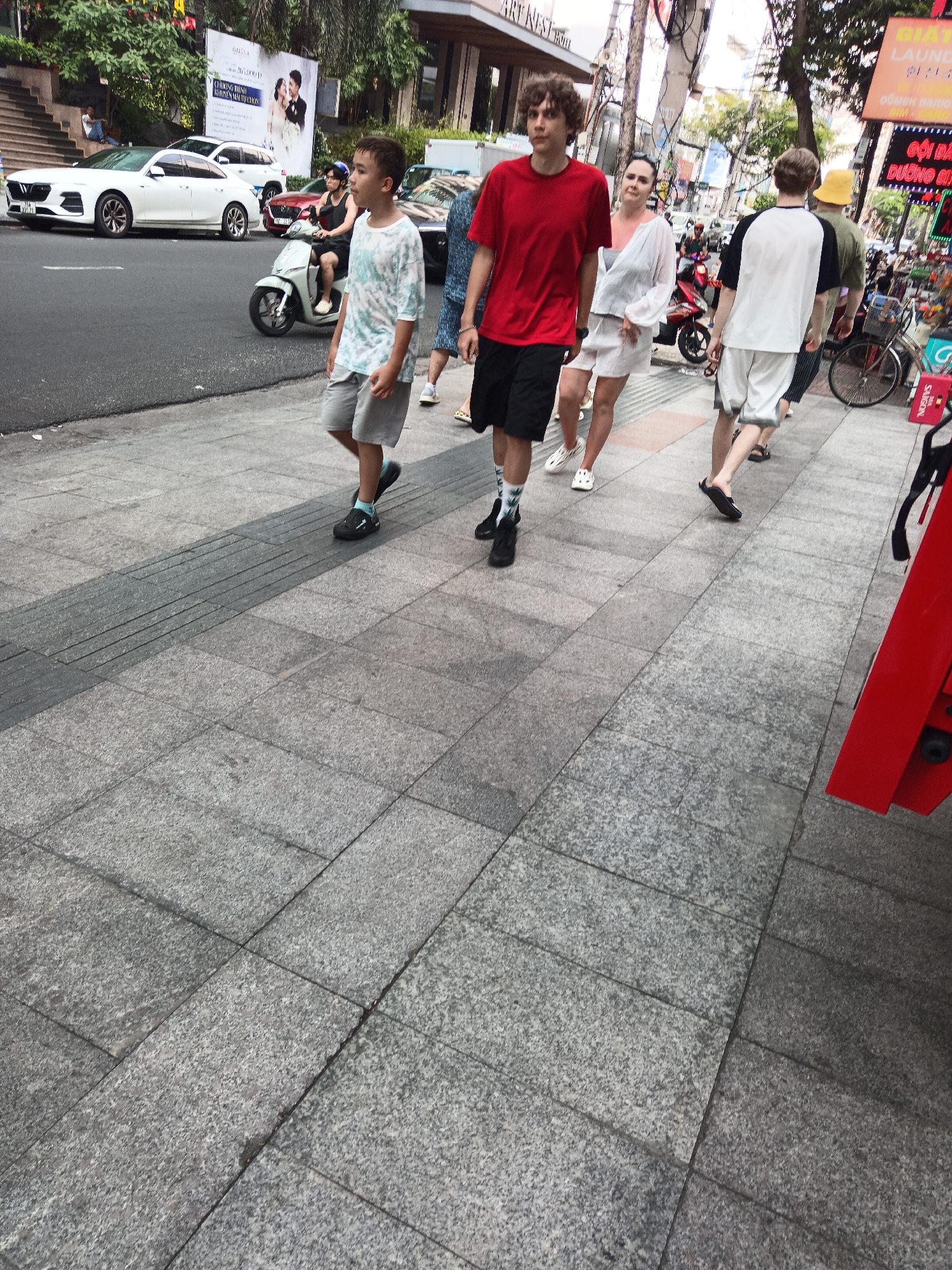 The image depicts a street scene in Nha Trang, Vietnam, with several people walking on a stone-tiled sidewalk. In the foreground, a young boy in a t-shirt and shorts is walking to the right. Behind him, a man in a red t-shirt and black shorts is also walking to the right. To the left of the man in the red t-shirt, a woman in a white top and shorts is walking. Further in the background, other individuals are visible, including a person wearing a yellow hat. Several vehicles, including a white car and a scooter, are present on the street. The environment includes buildings and trees. The lighting suggests it is daytime.