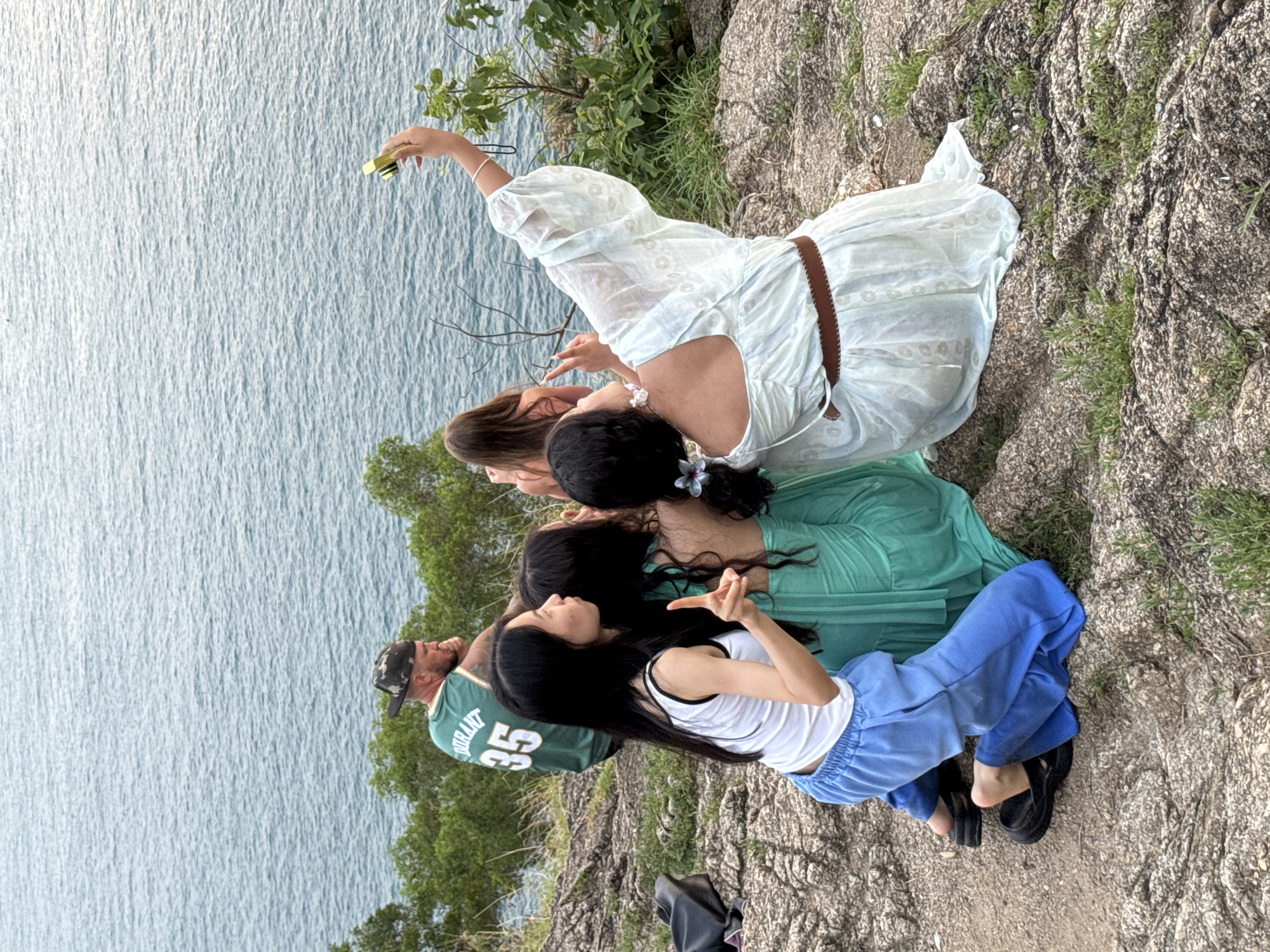 In Rawai, Thailand, four individuals are situated on a rocky, elevated outcrop overlooking a body of water. In the foreground, three young women are positioned close together, facing away from the camera. The woman on the right, wearing a light blue dress and a brown belt, is holding a smartphone aloft, taking a selfie. She has flowers in her hair. To her left, two women in matching teal dresses are also partially visible. One of these women is pointing her index finger upwards. In the mid-ground, a man wearing a green basketball jersey with the number 35, a camouflage baseball cap, and a beard is seated. Another young woman with long brown hair is partially visible in the background, also facing away from the camera. The environment consists of rocks, sparse vegetation, and the expansive blue sea and sky. A black bag is situated on the rocks to the left of the group.