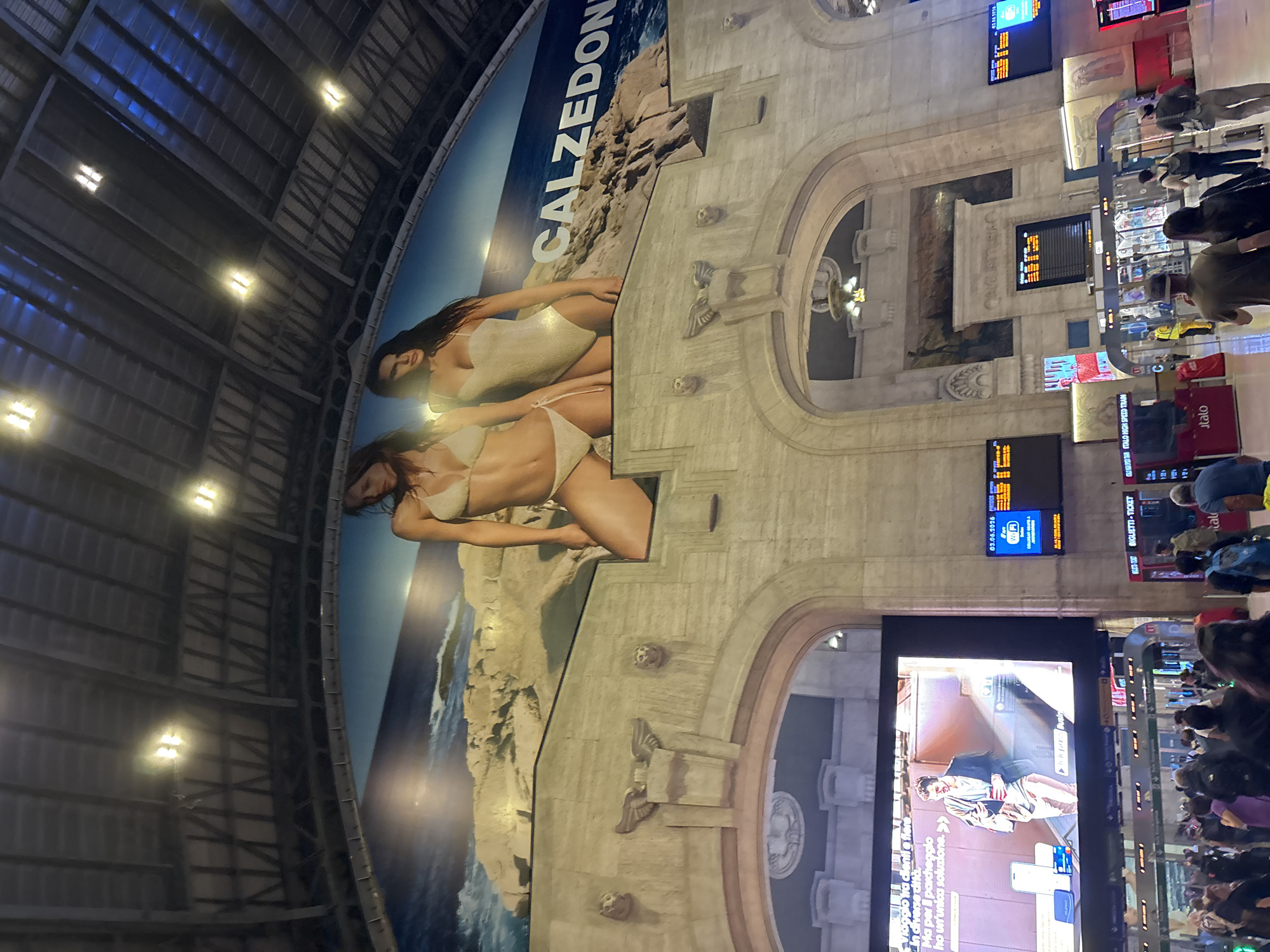 The image depicts the grand, high-ceilinged interior of what appears to be a major train station in Milan, Italy. The setting combines classic, ornate stone architecture with modern elements.
A colossal, curved advertisement for "CALZEDON" dominates the left side of the frame, featuring two women in swimsuits against a picturesque backdrop of a rocky coastline and the sea. The station's structure features intricately carved stone walls, arches, and decorative elements, including winged figures and sculpted faces, suggesting a historical and impressive building.
The upper portion reveals a vaulted ceiling with a metallic, industrial framework, interspersed with numerous bright, circular lights. Several large digital display screens are integrated into the stone architecture, showing various information. One screen prominently displays "03.06.2026," while others feature Italian text, such as an advertisement reading "Viaggio tra cientie... Ma per parcheggio ho un'unica soluzione.>>>"
In the lower right, a bustling crowd of people, including travelers with luggage, is visible, gathered near kiosks and information points. Clear signage, including "BIGLIETTI - TICKET" and "ITALO HIGH SPEED TRAIN," further confirms the location as a busy railway hub. The scene captures the dynamic atmosphere of a large public transport facility, blending historic grandeur with contemporary commercial displays and the constant flow of passengers.
