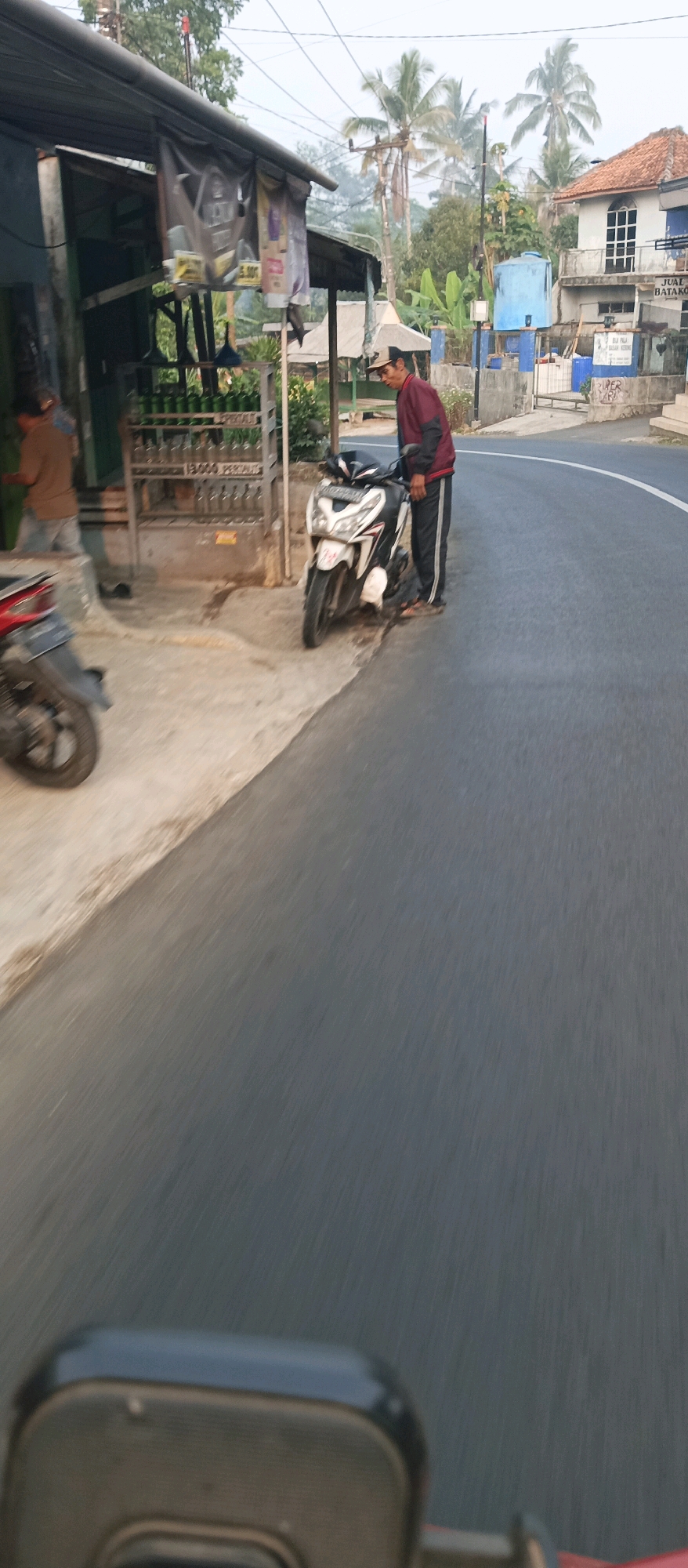 The image depicts a street scene in Purwakarta, Indonesia, likely during daytime given the lighting. A man wearing a maroon jacket and dark pants with white stripes stands next to a white and black motorcycle parked on the side of the road. The motorcycle is positioned on the paved street, which curves to the right. To the left of the man and motorcycle, there is a small roadside stall with a metal rack displaying multiple bottles filled with a light-colored liquid, presumably fuel, as indicated by signage that mentions "PERTALITE" and prices. In the background, there are buildings, including a white two-story house with a red tiled roof, and palm trees, typical of the tropical climate. Power lines are visible overhead, and a light blue water tank is also present. Another motorcycle, partially visible in the foreground, is parked on the concrete side of the road. A person in a light brown shirt and blue jeans is standing near the stall, interacting with it. The overall atmosphere suggests a rural or suburban setting along a road used for local commerce.