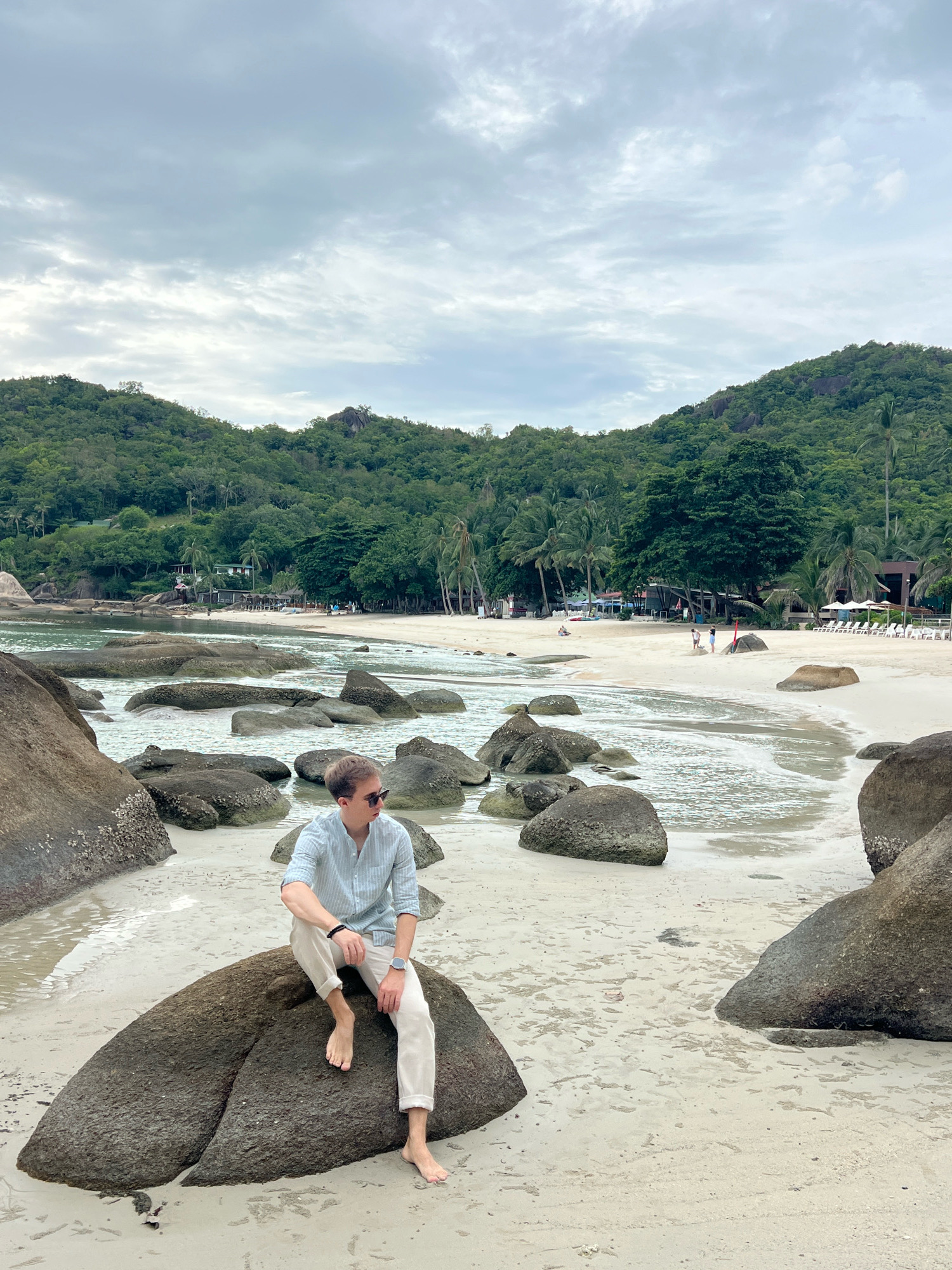 A young man with blonde hair, wearing a light blue striped button-down shirt, beige pants, and sunglasses, sits on a large rock on a sandy beach. He is barefoot and looking to his left. The beach is lined with palm trees and lush green hills in the background. The ocean is calm with several large rocks scattered in the water. The sky is overcast. The location appears to be a tropical beach in Ko Samui District, Thailand.