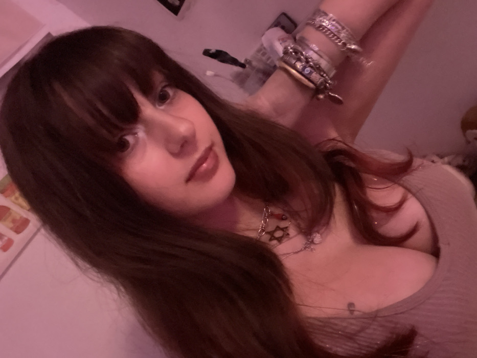 A woman with long brown hair and bangs poses for a selfie. She wears a beige top, silver necklaces, and a stack of bracelets on her raised arm. Her skin is fair, and she has glossy lips. The background includes a white wall with framed art, and hints of other objects are visible. The lighting gives the image a soft, warm tone.