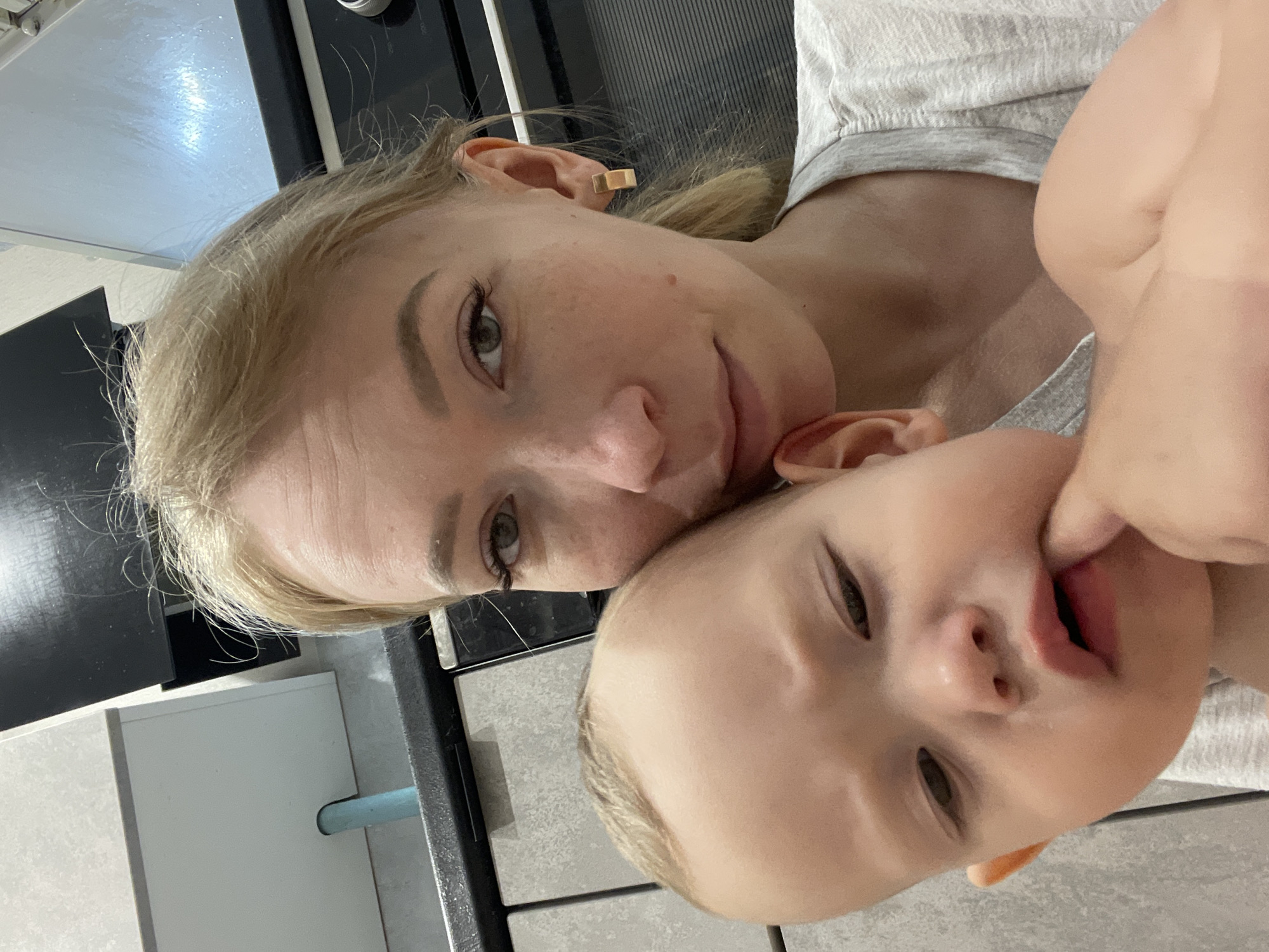 A selfie photograph taken indoors depicts a fair-skinned adult female with blond hair and a fair-skinned baby, possibly in Perm, Russia, based on the user's location. The woman is wearing a gold rectangular earring in her left ear. The baby has blond hair and appears to be sucking on their fingers. Both are wearing light grey clothing. The background shows a kitchen counter with a black stovetop and white cabinets.