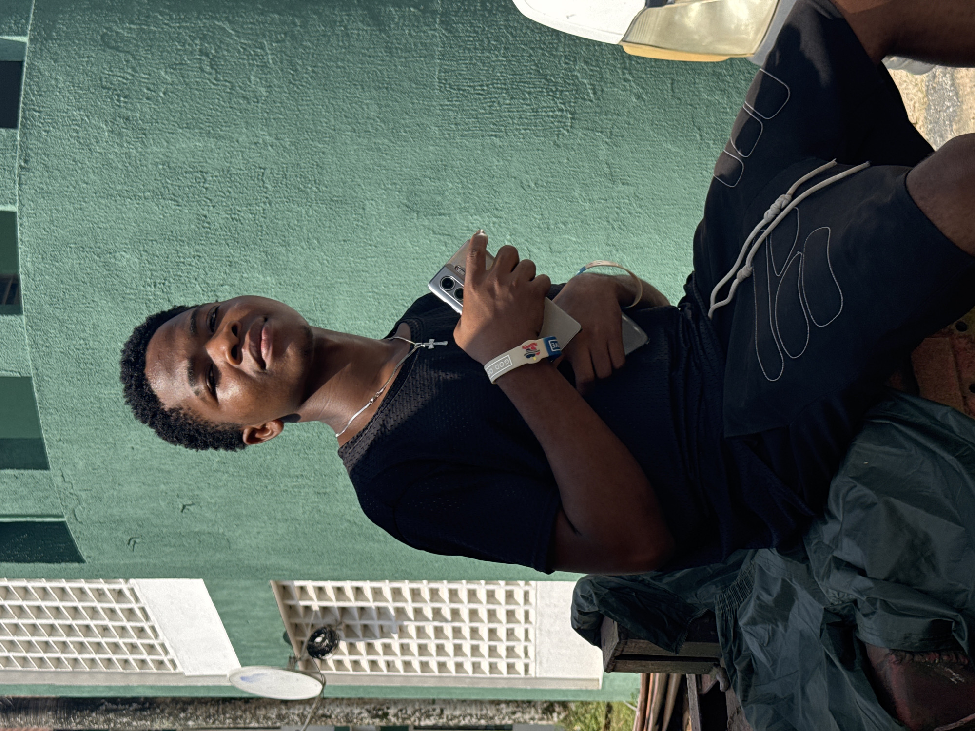 A young male, appearing to be an adolescent or young adult, is captured in an outdoor setting, looking directly at the camera with a neutral to slightly smirking expression. He has short, dark, curly hair and is wearing a black short-sleeved t-shirt and black shorts with white abstract, flame-like designs and white drawstrings. A silver-colored chain with a cross pendant is visible around his neck. On his left wrist, he wears two wristbands: a white one displaying the partial text "GOD OF" and a blue one showing "OVE." He holds a silver smartphone in his left hand and another dark phone in his right hand.
The scene is illuminated by bright natural light, indicating daytime with clear weather, likely morning or afternoon. The primary background element is a textured, light green (teal) wall. To the upper right, a portion of a white vehicle, including its headlight and bumper, is visible. To the lower left, in the background, there are structures featuring white, patterned ventilation blocks and a small, white, dish-shaped object. The subject appears to be sitting or leaning, with dark, crumpled fabric and some wooden elements beneath him. The location context for this image is Ihiagwa, Nigeria.