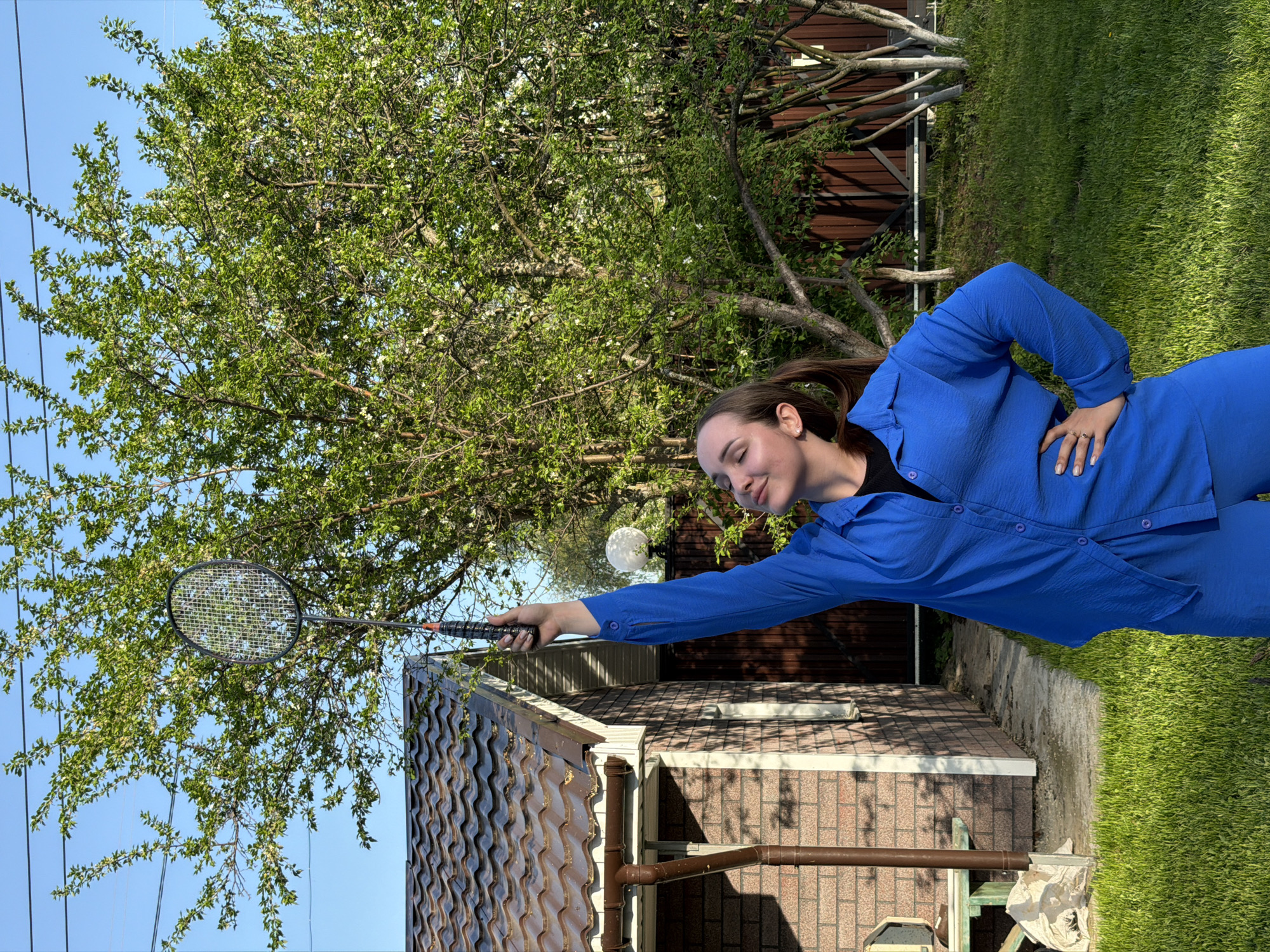 The image shows a young woman in a bright blue outfit standing outdoors, possibly in a backyard. She holds a badminton racket with her right hand, reaching toward a flowering tree. A single-story brick and tile roofed building is behind her to her left, and a wooden fence is behind the tree. There is green grass and foliage present, suggesting a residential area. The sky is bright blue. The image was taken in Chaikovsky, Russia.