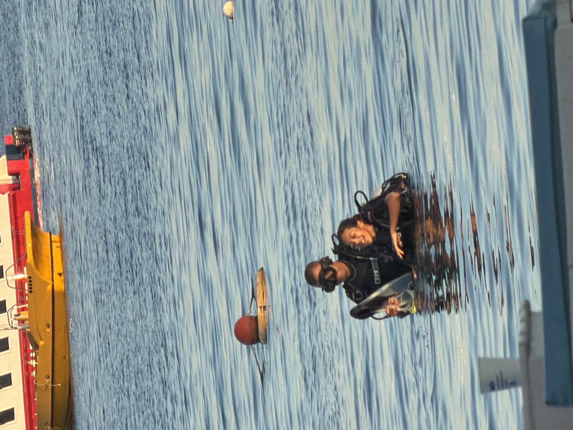 The image shows two individuals in scuba gear in the water. One is a male adult wearing a black wetsuit and a diving mask. The other is a female, also in scuba gear, with a diving mask on. They are both partially submerged in blue water. In the background, there is a yellow boat with a red and white superstructure. A red and white buoy with a yellow float is visible in the water to the left of the divers. Another small white buoy is visible further out in the water to the right. The scene appears to be in a coastal area, likely the Red Sea given the location context provided. The water is calm with small ripples. The lighting suggests it is daytime. There are no specific landmarks or identifying features to pinpoint the exact location within Sharm El Sheikh, Egypt.