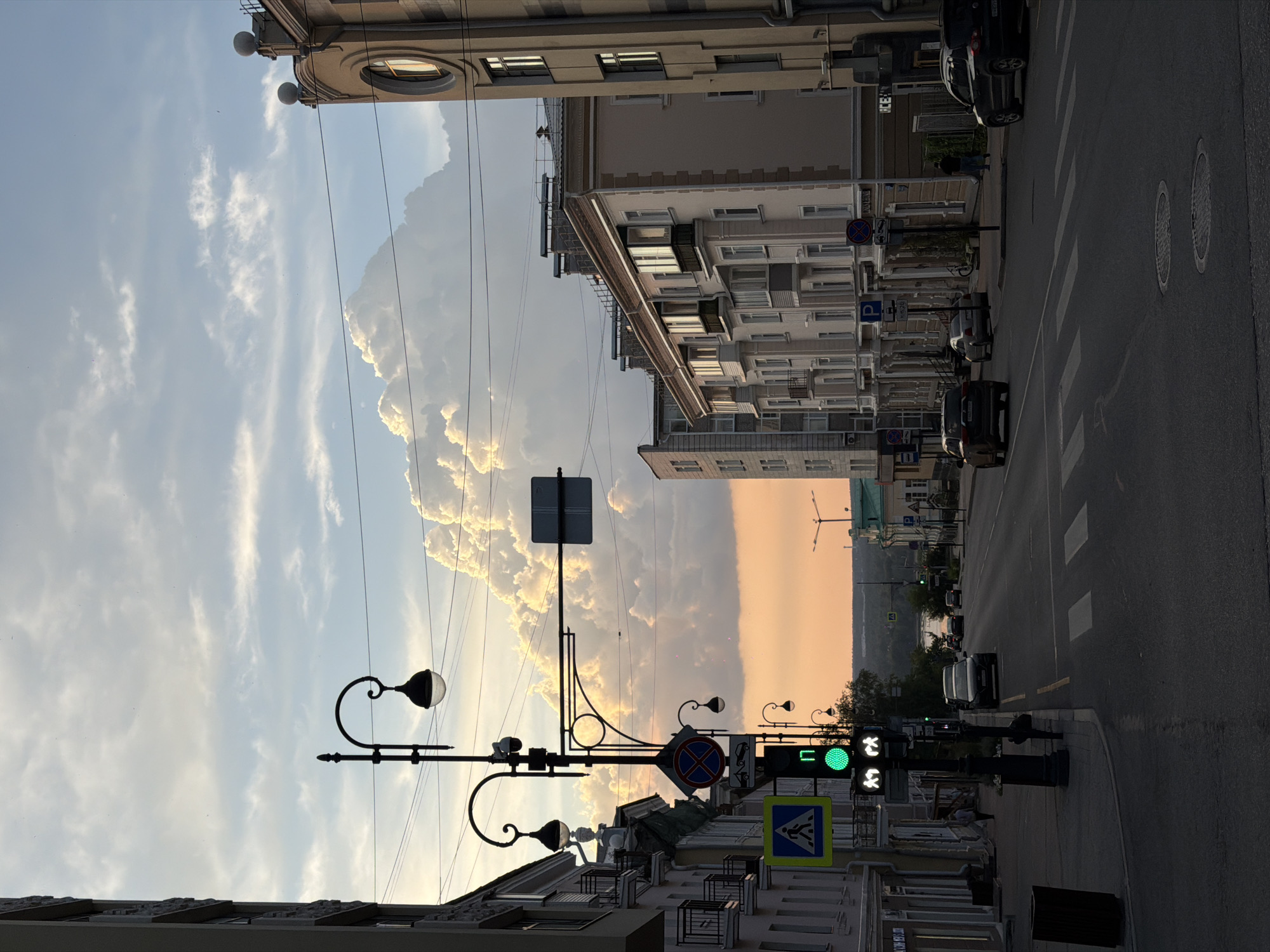 This is a street scene captured in Perm, Russia, during what appears to be late afternoon or early evening. The sky is dramatic, with large, puffy white clouds illuminated by the setting sun, casting a warm orange glow on the horizon. The city's architecture is visible, with multi-story buildings lining the street.
On the left side of the frame, a tall, slender building with a stone facade is partially visible, its dark windows reflecting the sky. Adjacent to it is a building with lighter colored walls, featuring balconies with decorative railings.
The street itself is paved and shows multiple lanes of traffic. A pedestrian crossing is marked on the road. Several cars are present: a dark sedan is driving away from the viewer in the right lane, a light-colored sedan is parked further down on the left, and another dark SUV is seen in the distance on the right.
A prominent traffic light signal for pedestrians is green, showing two walking figures. The traffic light for vehicles is also visible. Several street lamps with an old-fashioned design are mounted on poles. Road signs, including a pedestrian crossing sign and a "no stopping" sign, are also present.
On the right side of the street, a beige building with arched windows and decorative elements on its facade stands out. Further down, another building with green trim is visible.
A few individuals are sparsely scattered in the scene. One person is walking on the sidewalk on the left, and another individual is seen walking on the right side of the street.
The overall atmosphere suggests a typical urban environment at the end of the day, with the natural beauty of the sky contrasting with the man-made structures of the city.