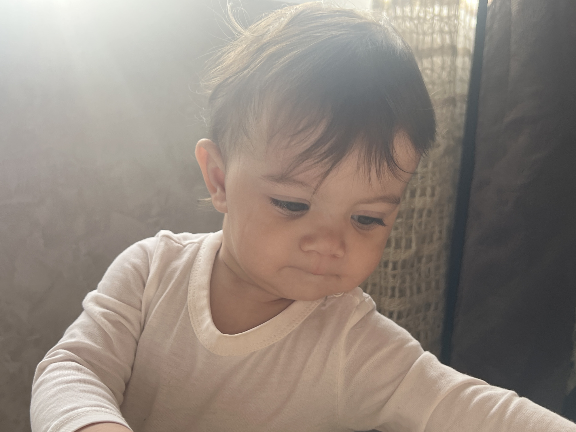 A light-skinned infant or toddler with dark brown hair is depicted from the upper chest upwards, positioned off-center in a vertical composition. The individual wears a plain, light-colored, long-sleeved garment with a rounded neckline. The subject's head is tilted slightly downwards and to the right, with eyes directed towards the lower right portion of the frame, and lips slightly pursed. Strong directional light emanates from the upper left, creating a significant lens flare and illuminating the subject's hair and left shoulder. The background is out of focus, featuring a light grey, textured wall to the left and a darker, undefined fabric or curtain to the right. The scene is located in Elena, Bulgaria.