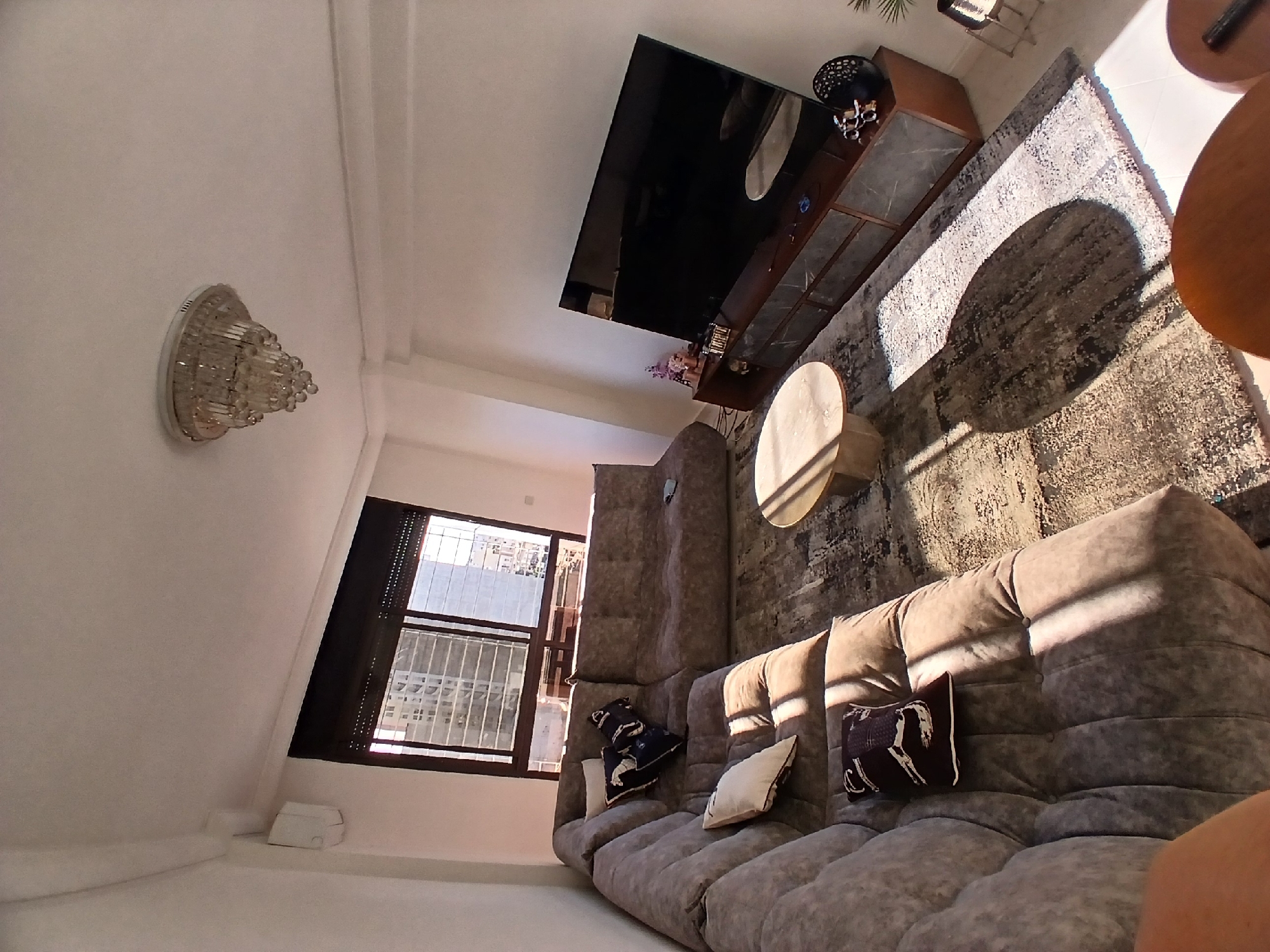Here is a view of a living room. A sectional sofa is placed in front of a window with metal bars. Pillows are on the sofa. A coffee table is next to the sofa. A television is mounted to the wall. A rug is on the floor. A light fixture is on the ceiling.