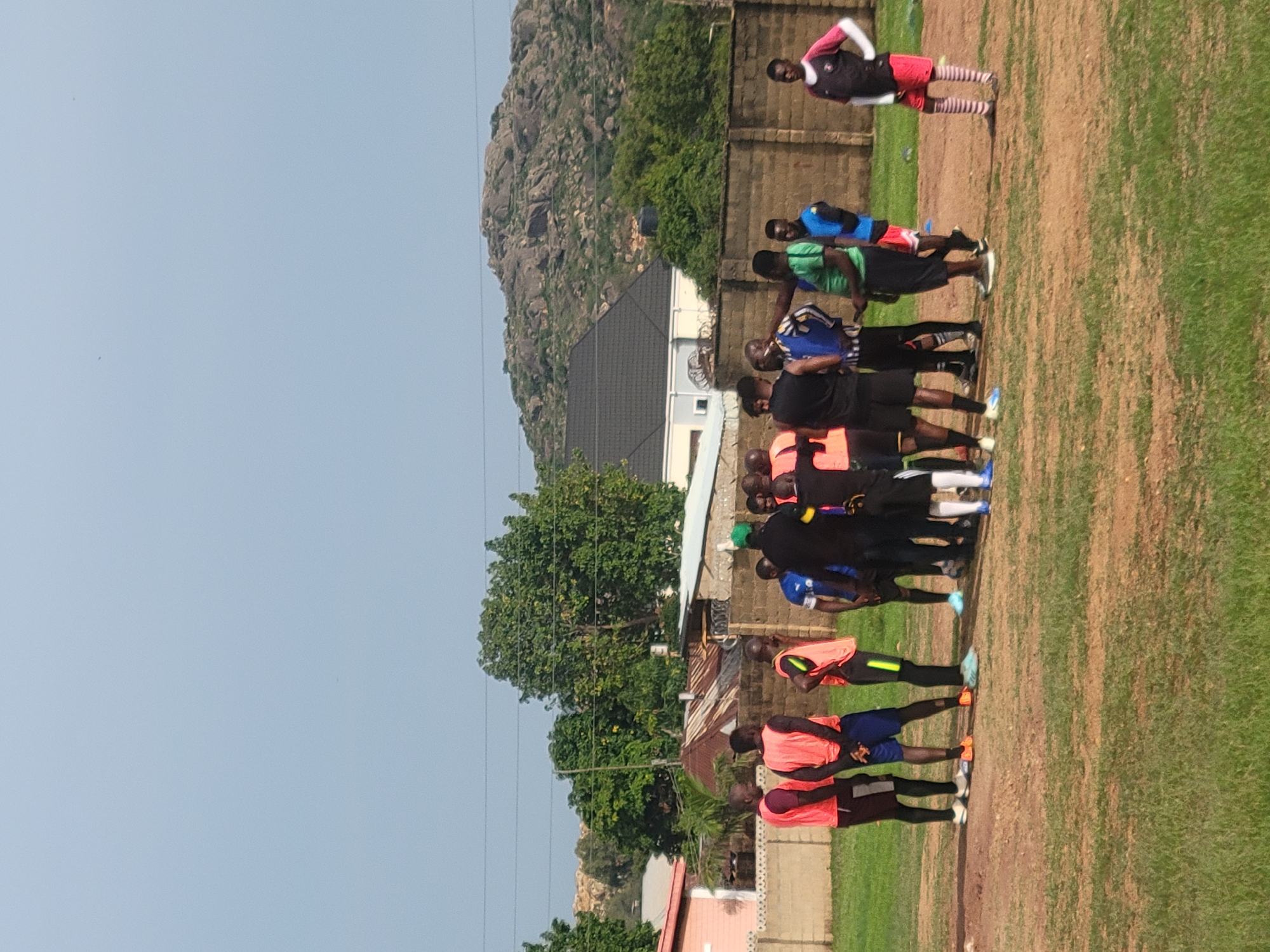 The image shows a group of approximately 15 men standing on a grassy field. They are wearing various sportswear, including jerseys, shorts, and socks of different colors. Some appear to be in a line formation, possibly stretching or warming up. In the background, there's a residential area with houses and trees. A prominent rocky hill or mountain can be seen in the distance. The location is Jos, Nigeria.