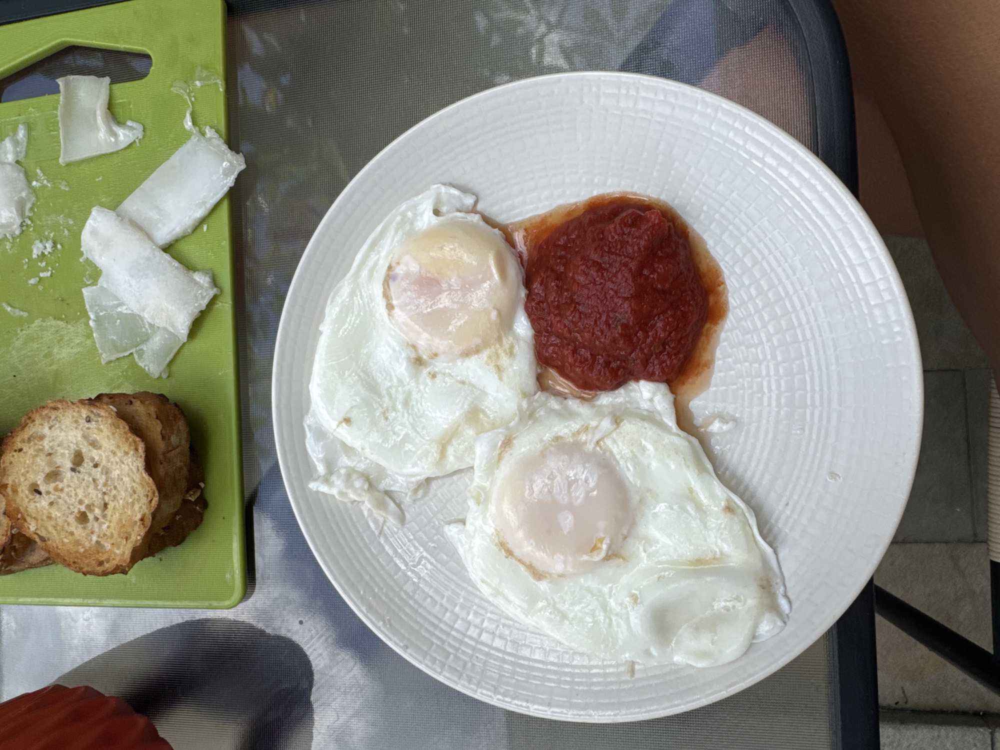 The image shows a meal served on a table. There are two fried eggs with runny yolks and a portion of red sauce, likely tomato-based, on a white plate. A green cutting board contains sliced white cheese and toasted slices of bread with visible seeds. A person's leg is partially visible in the background. The setting could be outdoors. I cannot verify the location to be Kuta Selatan, Indonesia from the given image.